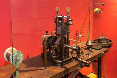 The metal knitting machine stands in the middle of a wooden workbench. There is a hand crank on the front. A white spindle stands to the left of the machine. A metal thread is fed through the machine’s two wheels, which are stacked one on top of the other.