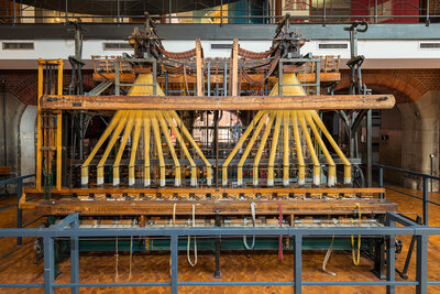 The loom is about five meters wide, 2.4 meters deep, and 3.5 meters high. The two loom attachments are suspended from a metal frame at the top. They look like two giant rakes, each with eight tines.