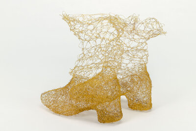 Two boots stand side by side. They are woven from fine copper wire that looks like a delicate gold thread. The material is woven more densely at the toes and heels. Toward the top, the material becomes more airy, as if the boots were dissolving.