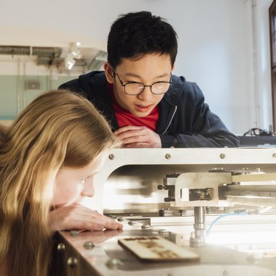A girl and a boy are looking at a large piece of machinery. The girl is leaning over the opening to get a better look inside, while the boy is leaning against the edge of the machine.