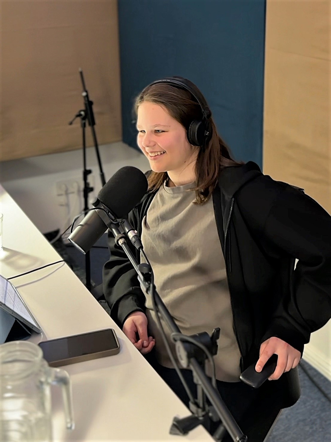 A young person with shoulder-length brown hair sits in a recording studio in front of a microphone and smiles.