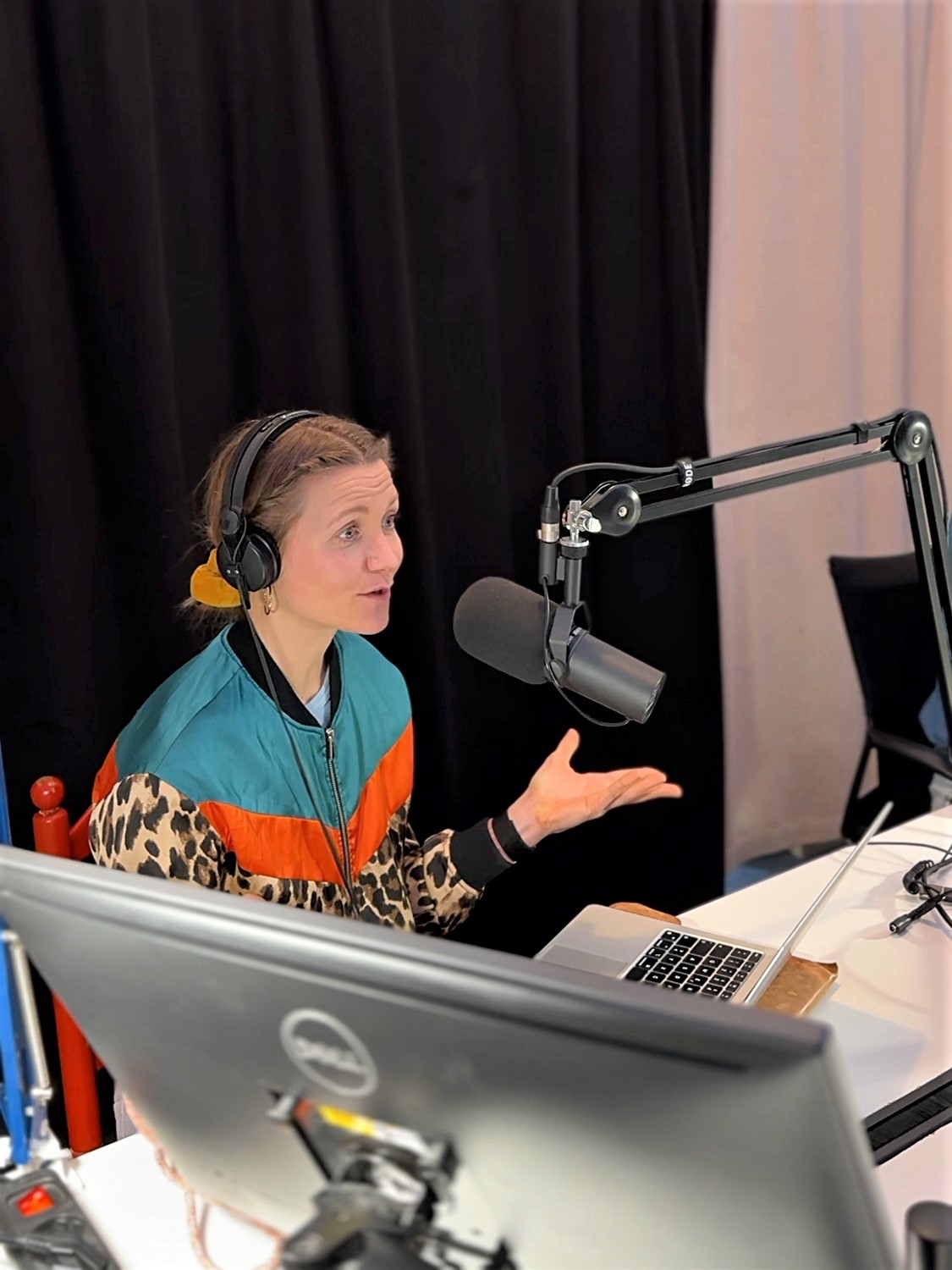 Host Jana Forkel sits at a table in a recording studio with a microphone and a laptop. She is wearing a colourful patterned jacket and speaking into the microphone.