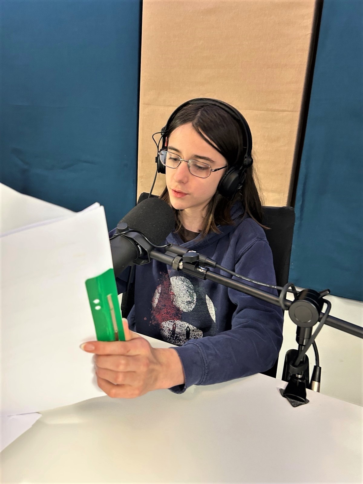 A young person with shoulder-length brown hair and glasses sits in a recording studio in front of a microphone, holding a script.