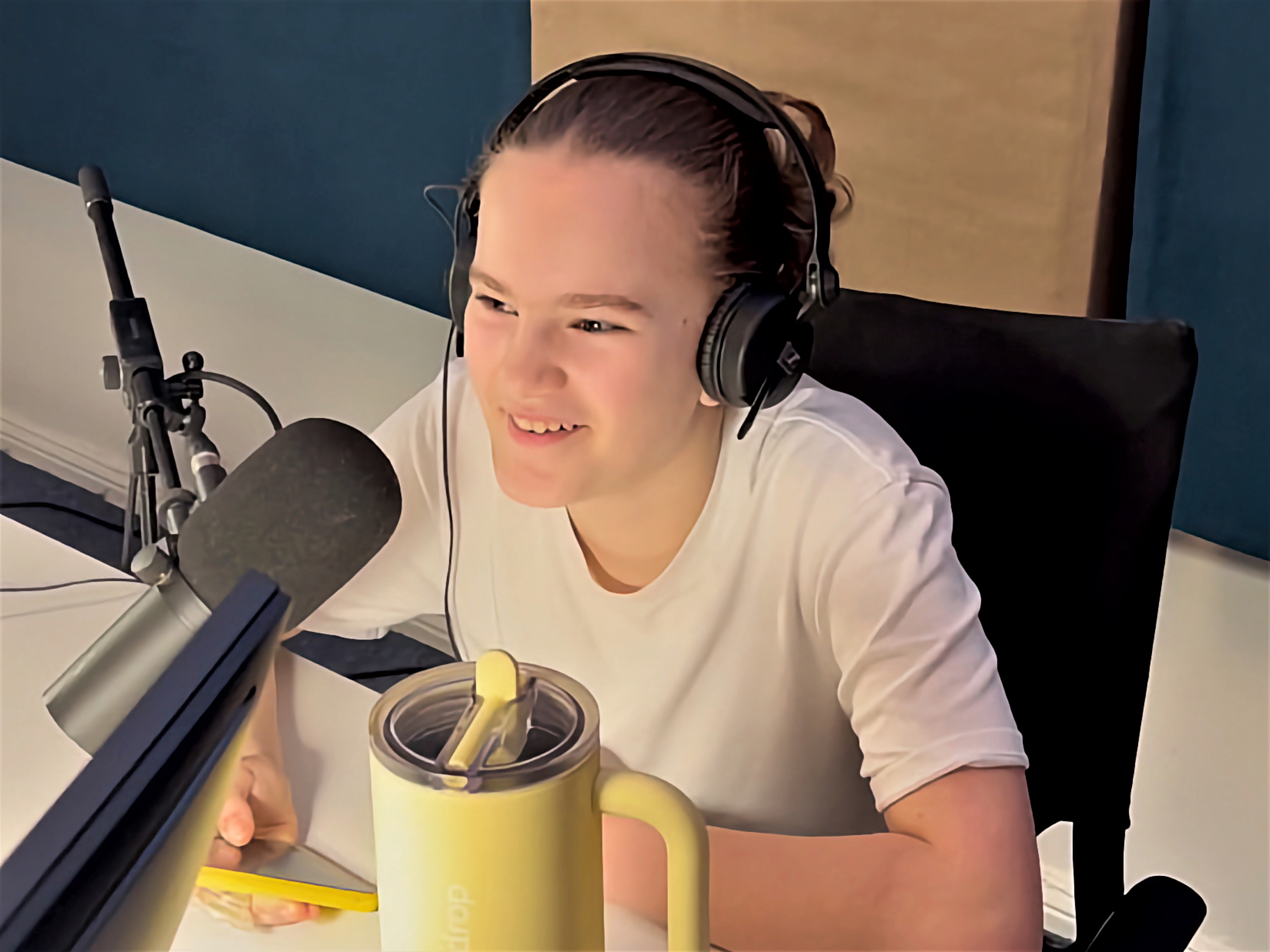 A young person with brown hair sits in a recording studio in front of a microphone and smiles. They hold a smartphone with a yellow case in their right hand.