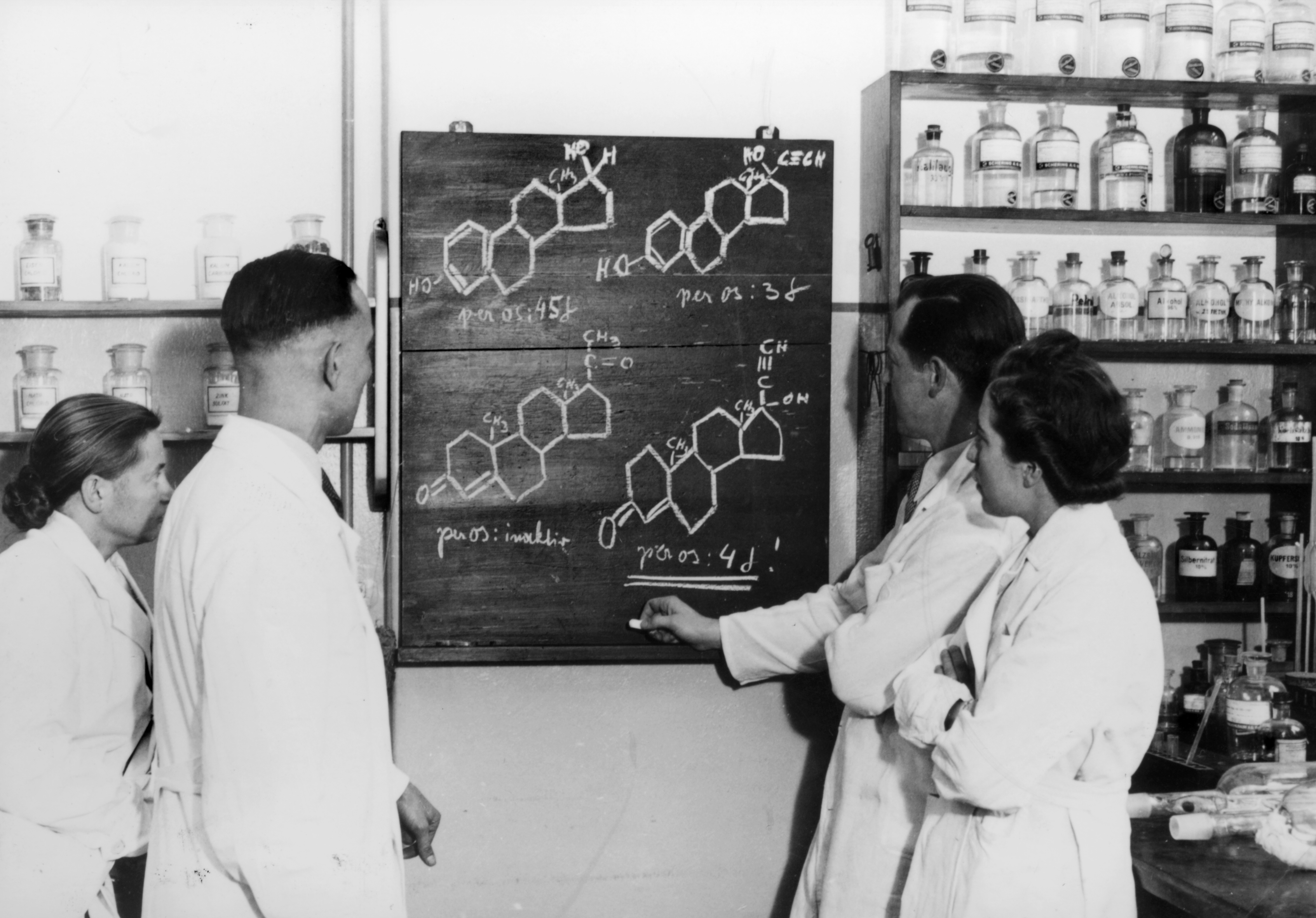 Black-and-white photograph, 1938: In the Schering company’s laboratory, two male employees and one female employee are standing in front of a blackboard. Molecular structural formulas have been drawn on it in chalk.