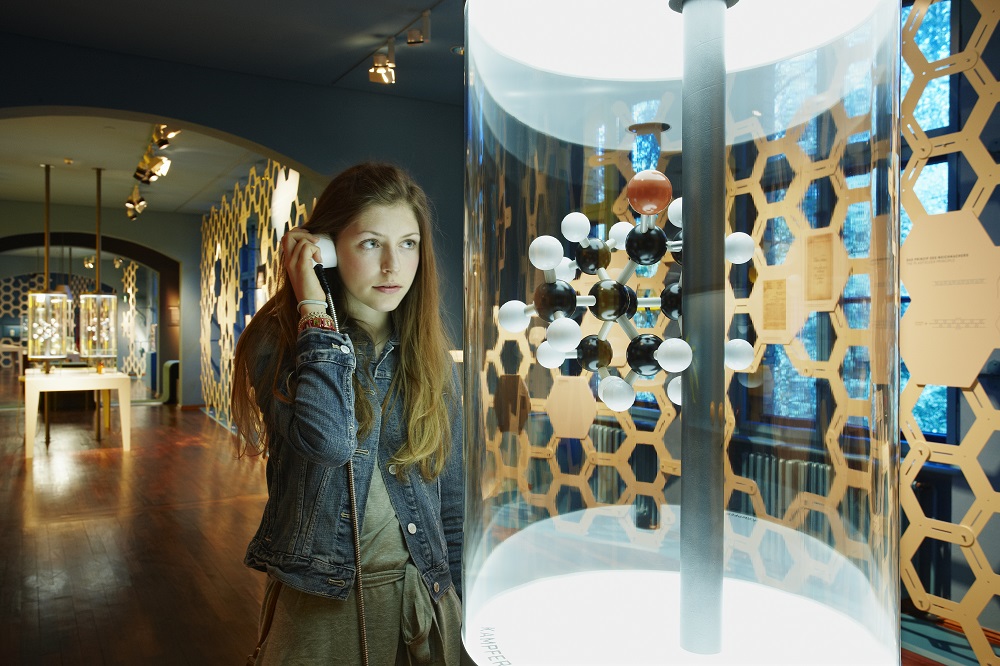 A visitor is holding the earpiece of an audio station to her ear. As she does so, she is looking at a model of a molecule made up of black, white and red spheres.