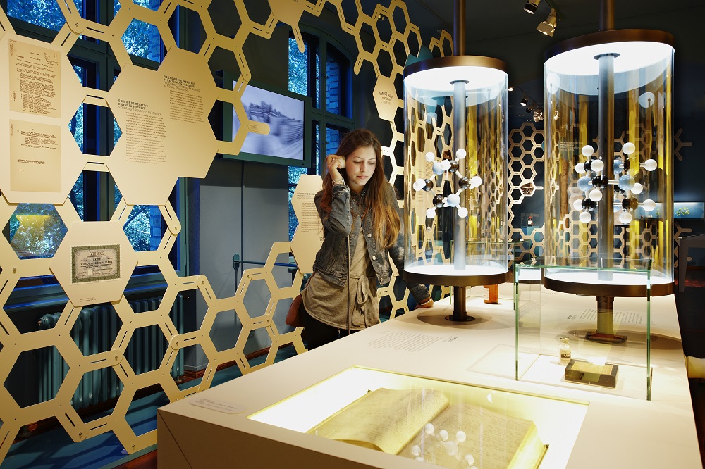 A visitor is standing at an audio station. She is looking at two molecular models made up of black, white and red spheres.
