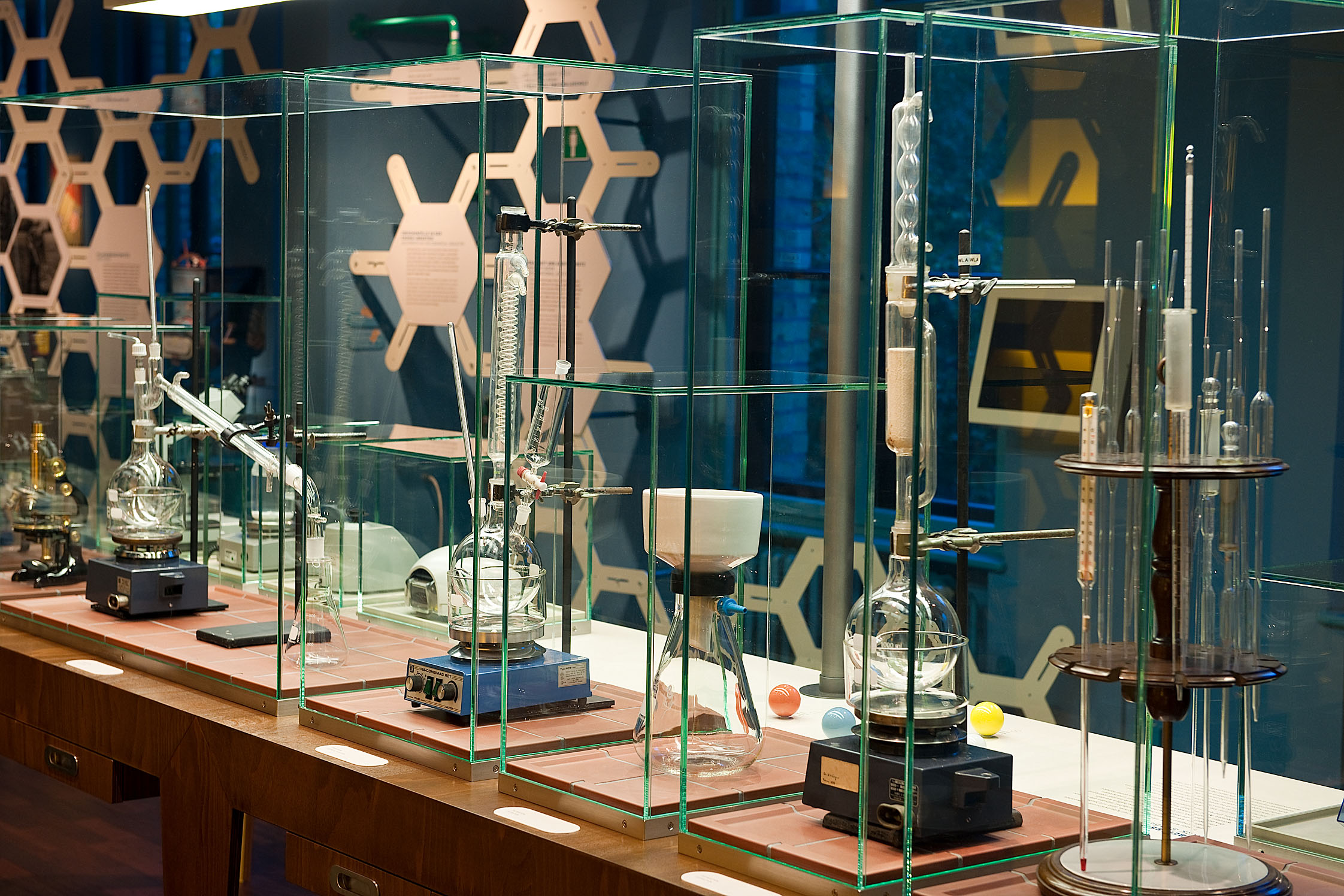  There are several display cases on a table. They contain various instruments from a chemistry laboratory, such as pipettes, a Liebig condenser for distillation, a centrifuge and a microscope.