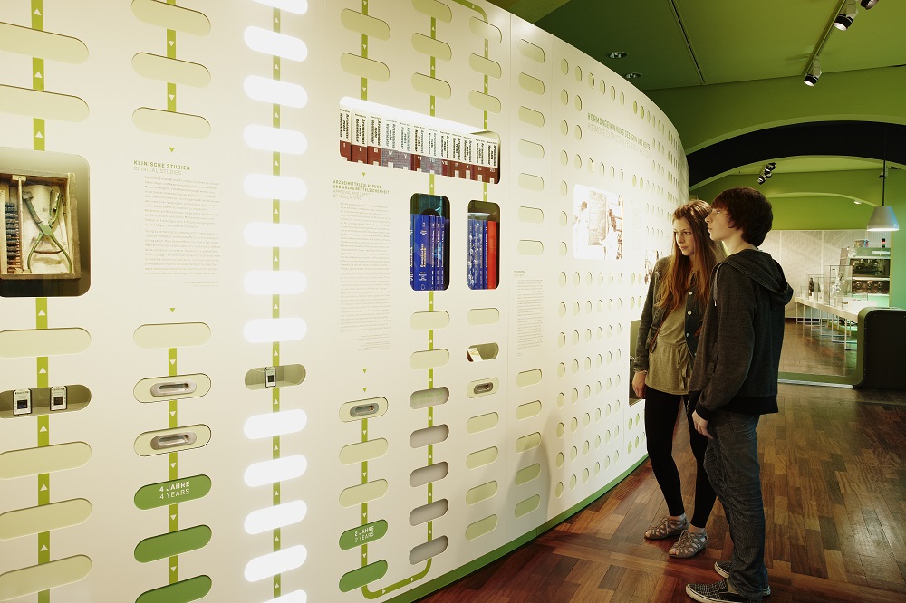 A female visitor and a male visitor are looking at an infographic which uses green and white light fields to illustrate the journey from the search for active ingredients to the finished medicine.