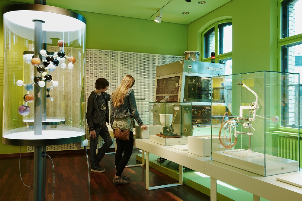 A female visitor and a male visitor are looking at the row of machines and instruments on display, which process chemical substances into tablets, pills, powders or ointments. Next to them is a model of the penicillin molecule.
