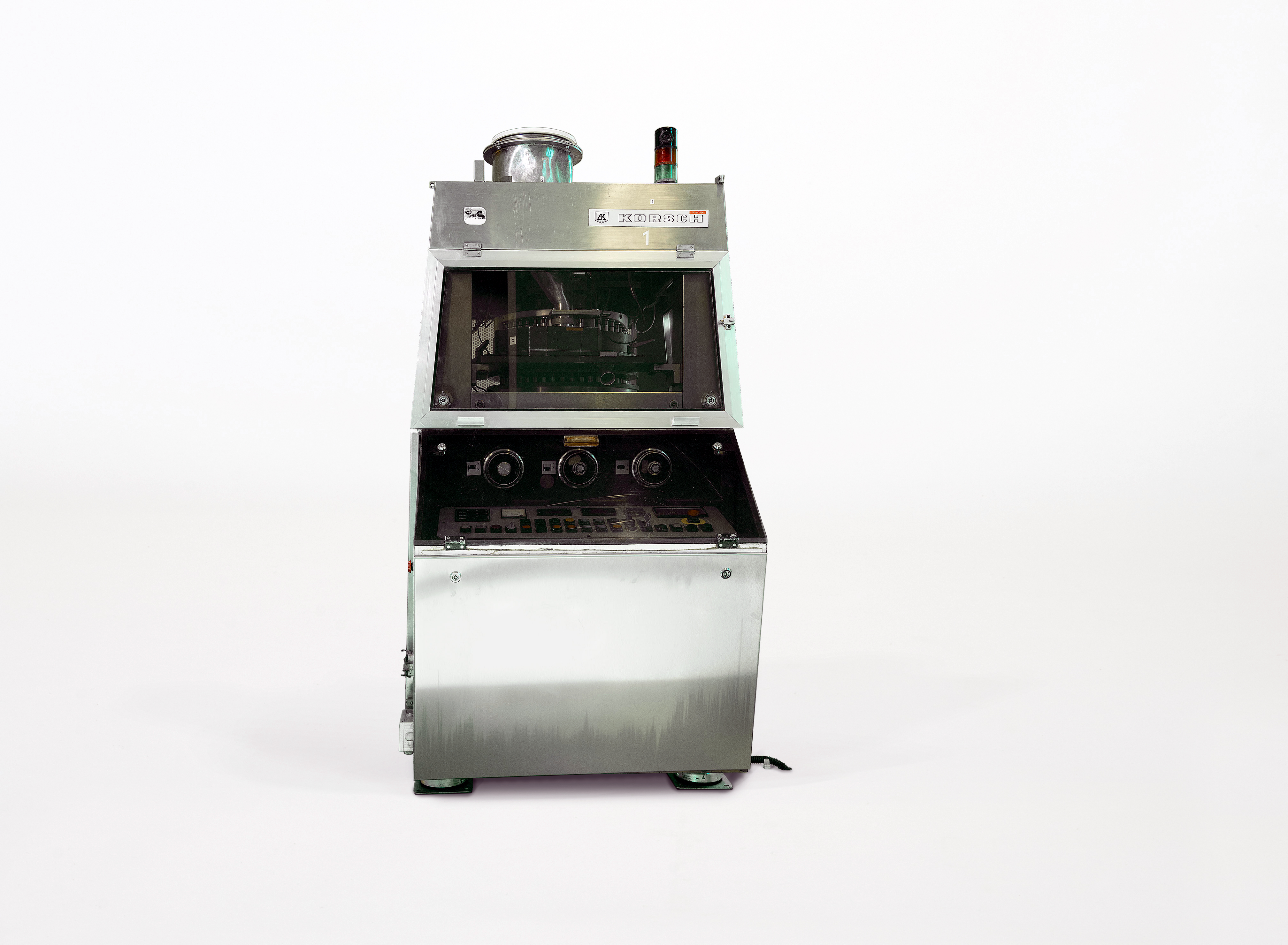 The tablet rotary press resembles a large slot machine. At the front is a protruding control panel with three large rotary switches and a keypad. There is a large viewing window at eye level.