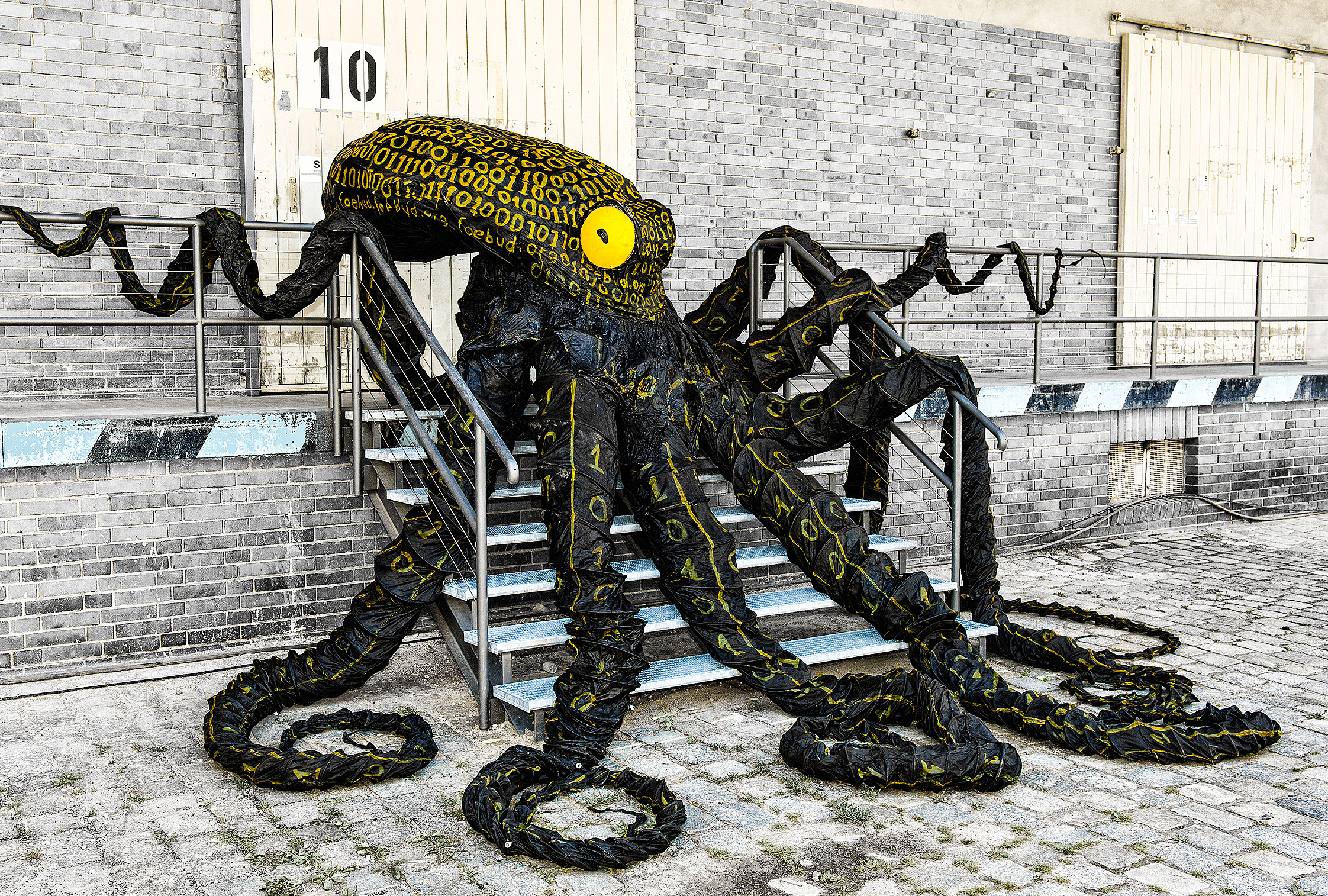 Otto the Data Octopus is lying on a wide metal staircase in front of a wooden gate. His eight black arms are draped across the steps. His eye glows yellow. His head and arms are covered with yellow zeros and ones.