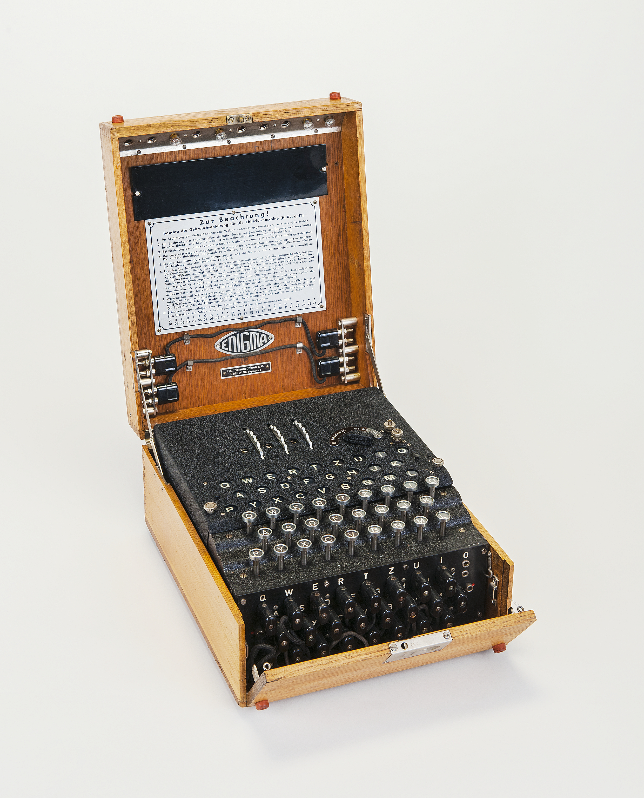 The black machine sits inside a wooden box that is open. On top, it has a letter keyboard, a letter display, and three dials. On the front of the machine are black two-pin jacks.