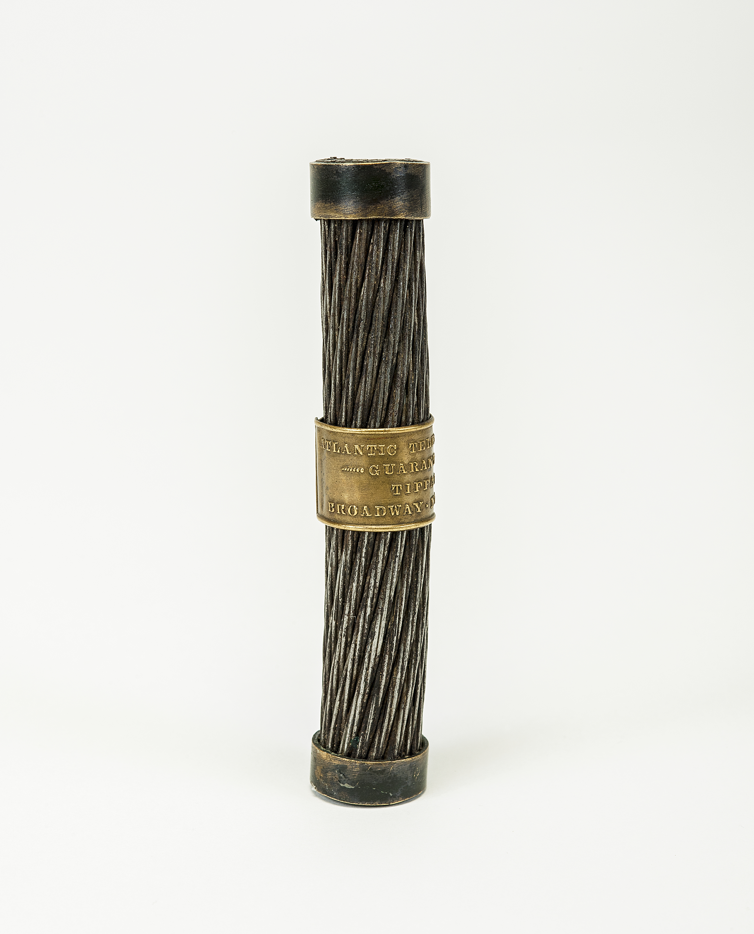 The section of the transatlantic cable stands upright. It has a metallic sheen. The individual cable strands are visible. In the center is a brass-colored metal band with an inscription in raised letters.