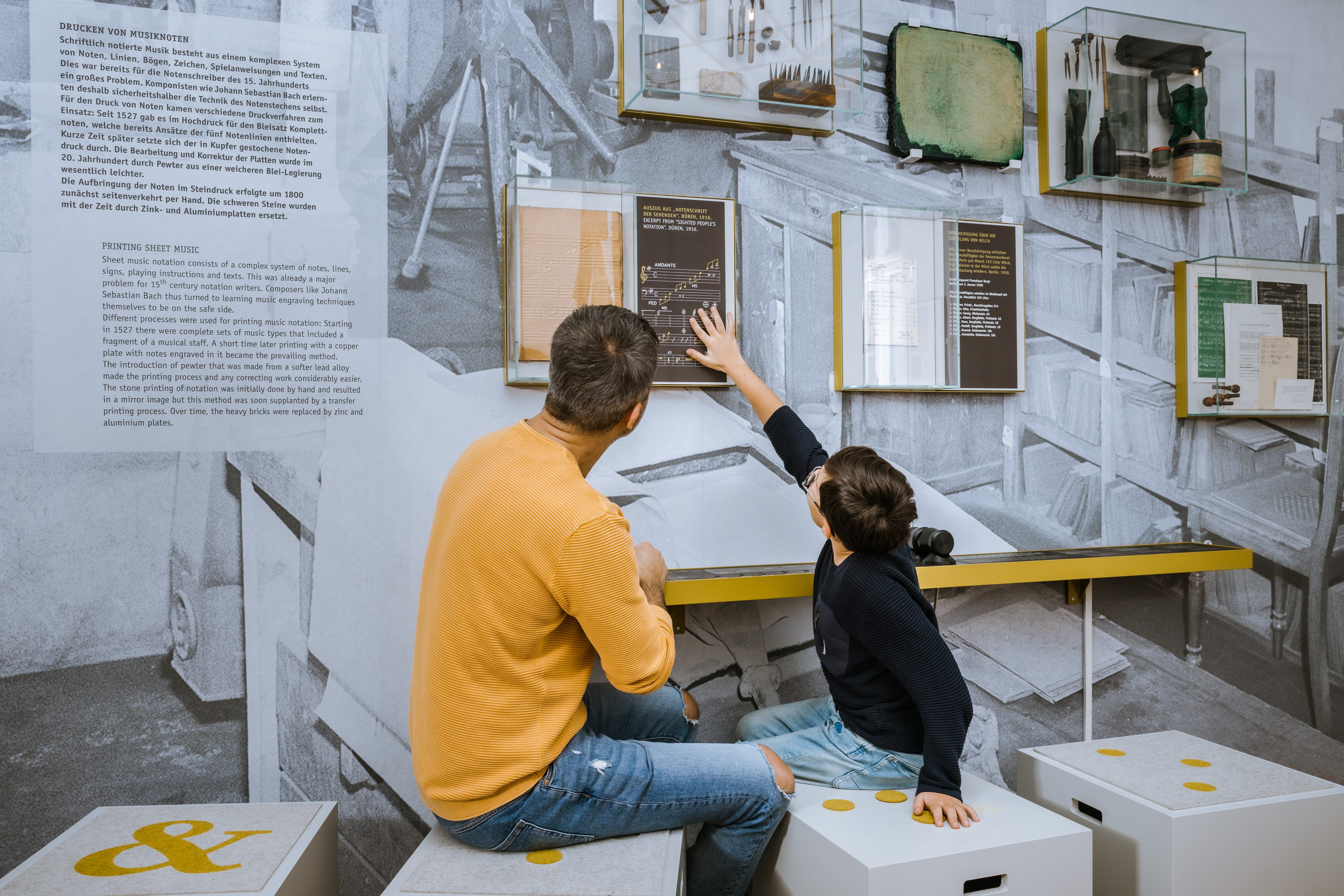 A man and a boy are sitting in front of a display wall with printed sheet music. There is also a tactile object on the wall, which the little boy is touching.