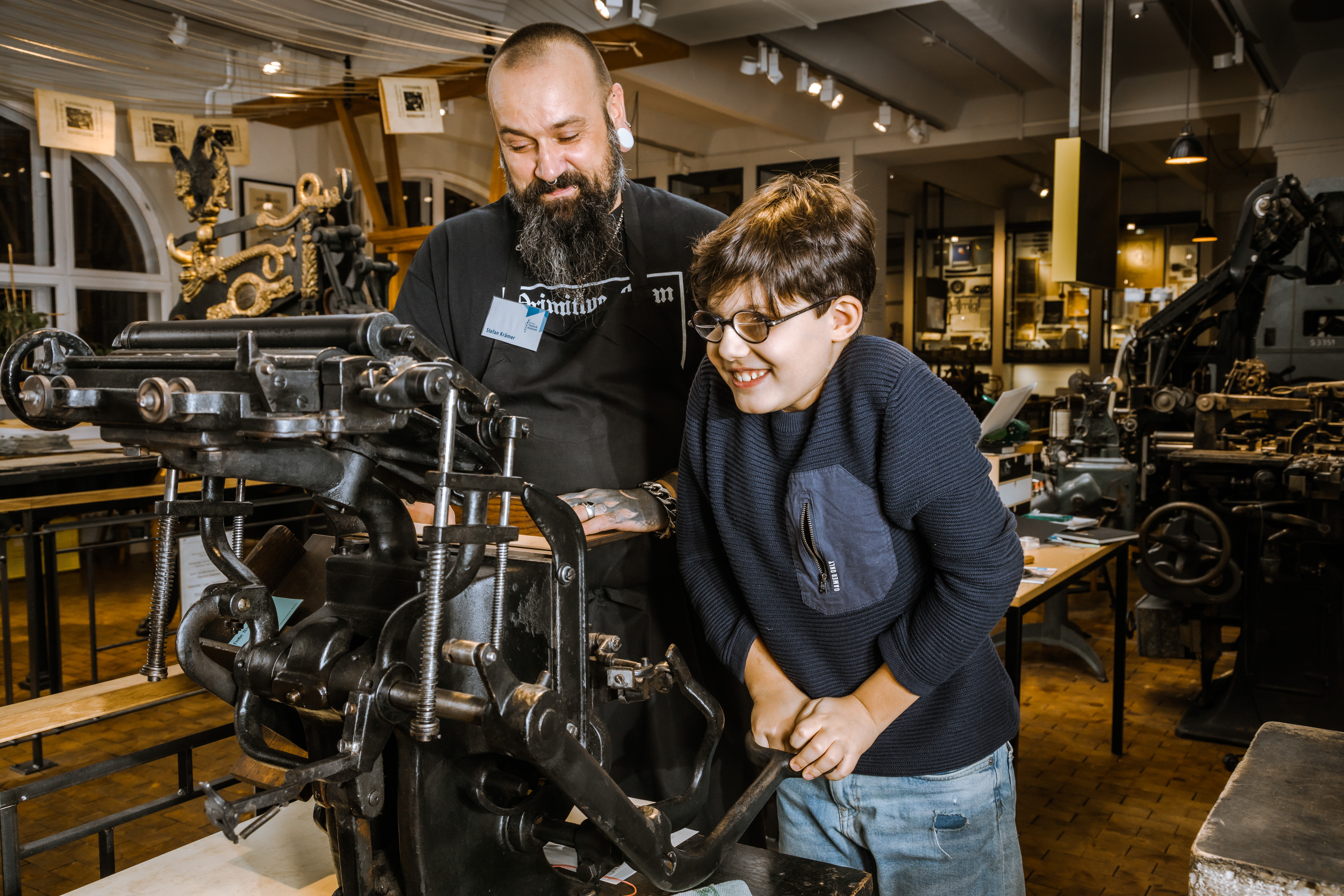 A little boy is operating a printing press; a bearded man is standing next to him, helping him.