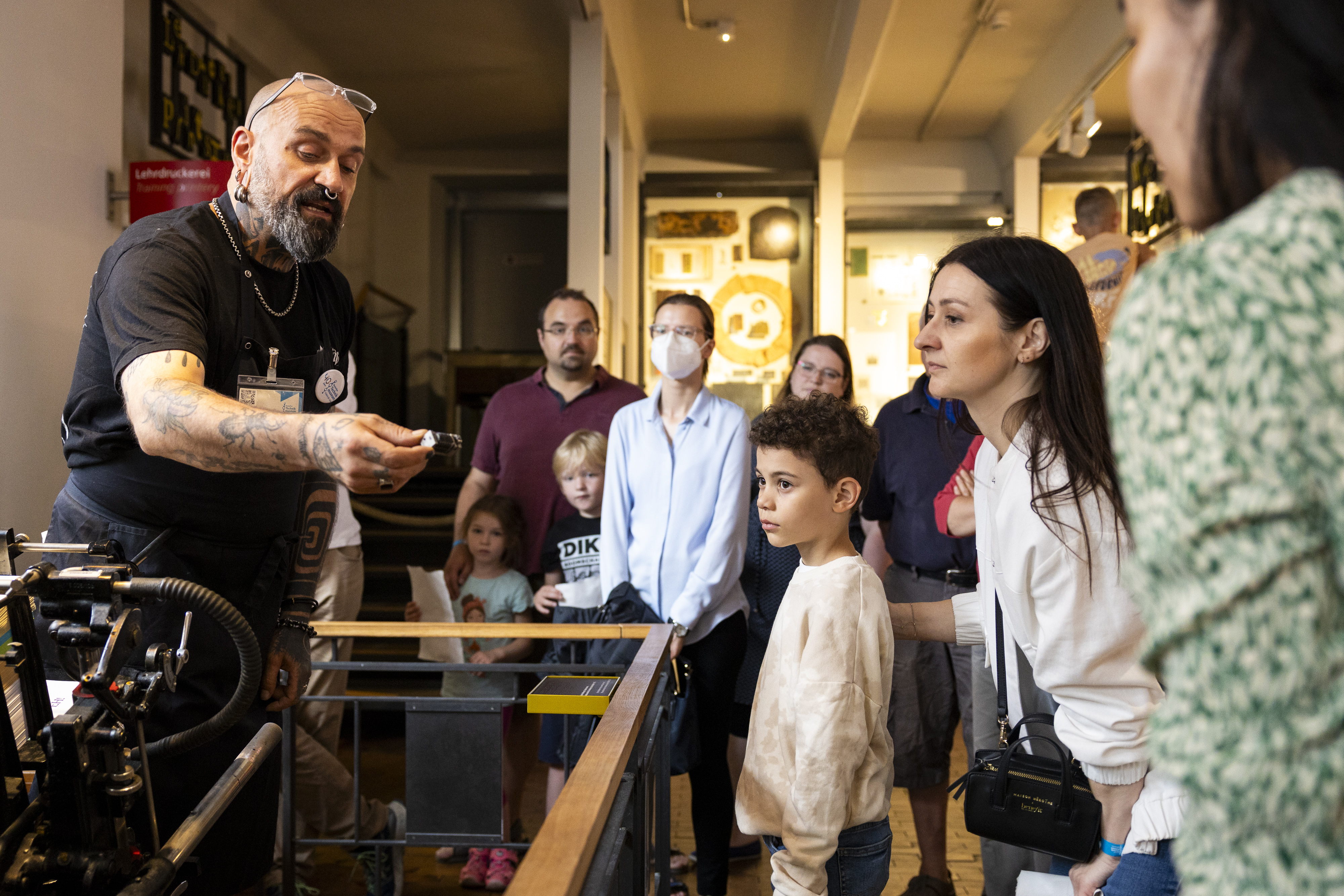 A man with a beard is standing at a printing press, demonstrating how it works to a group of adults and children.