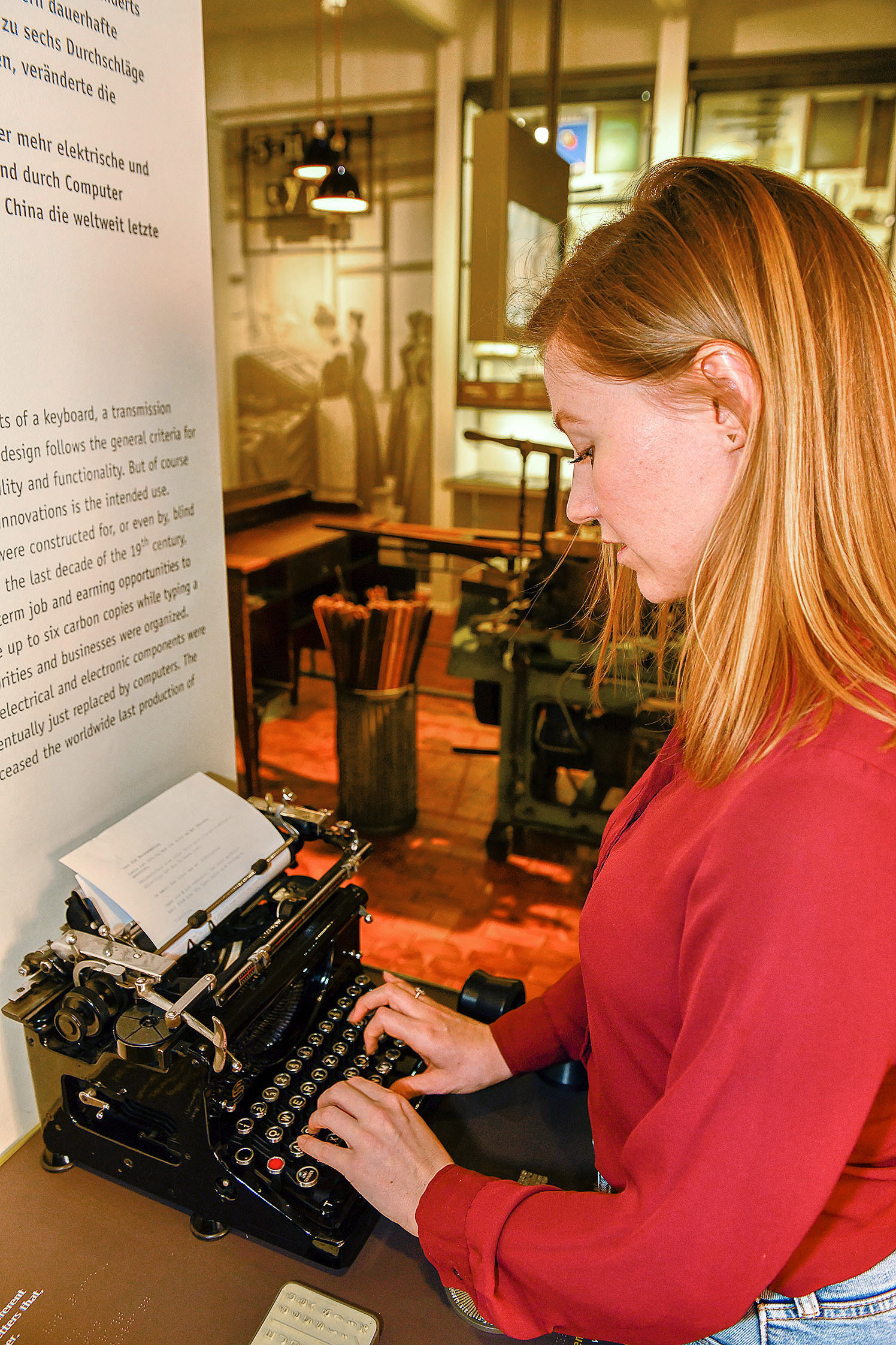 A young woman is standing in front of a black typewriter with a sheet of white paper loaded in it. Her fingers are tapping on the keyboard.