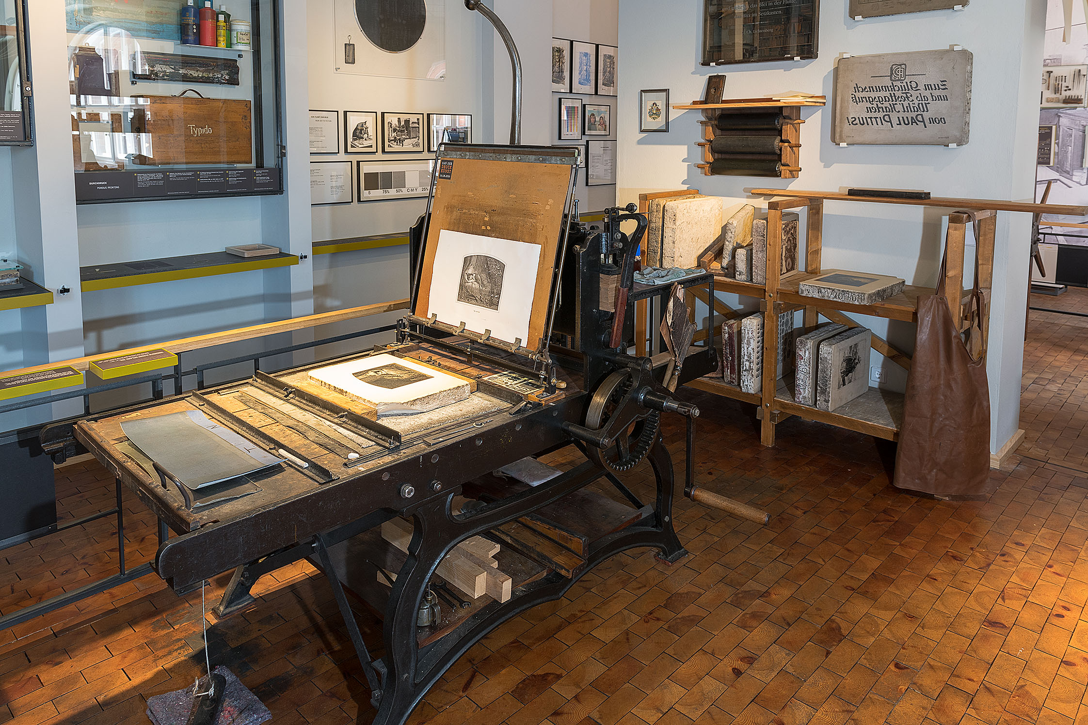 A light-colored, polished lithographic stone lies in a large, iron lithographic press. A white sheet of paper is stretched across the open brown lid of the press, already showing the finished print of the design.