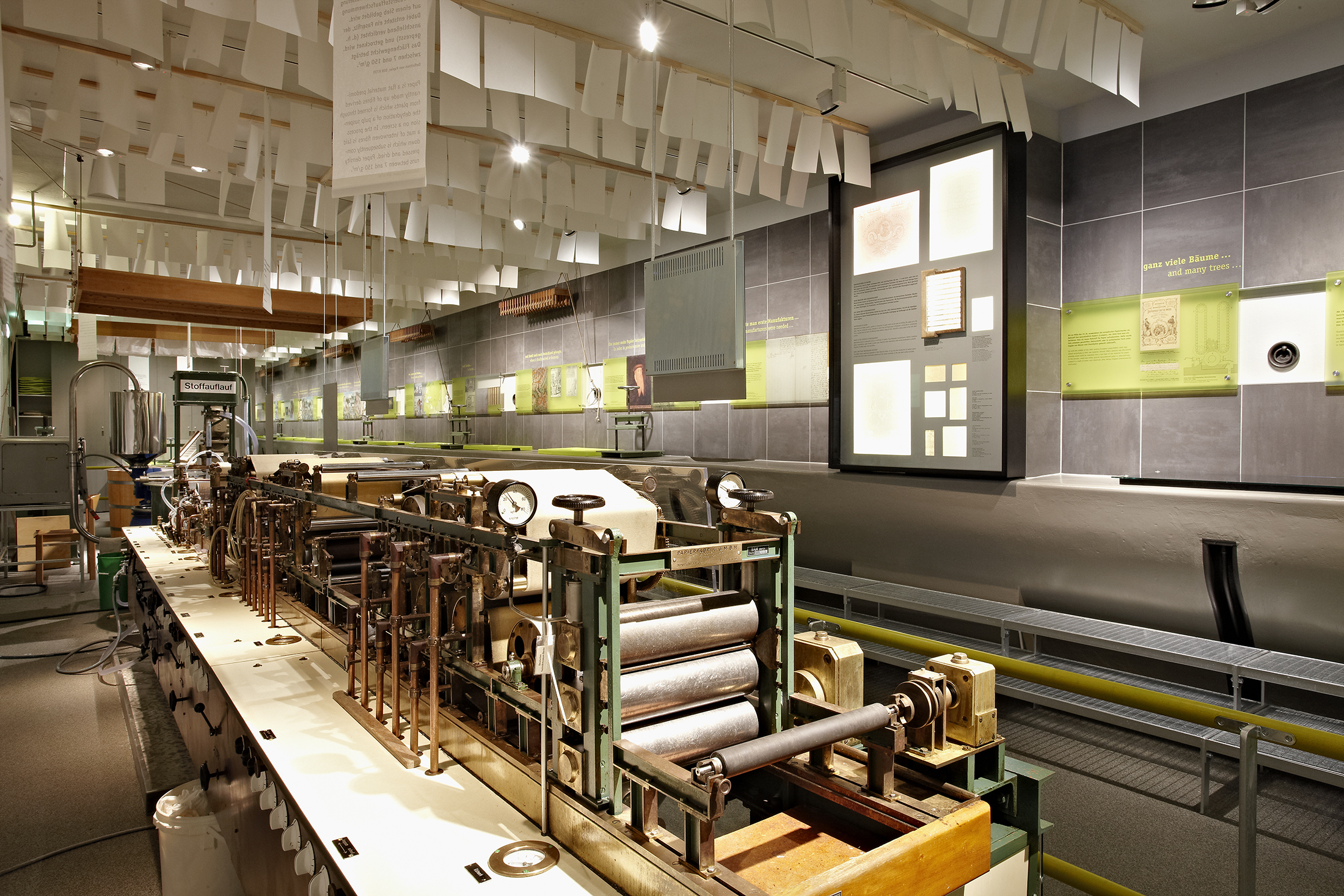 A view of the "Papermaking" exhibition. In the foreground is a machine several meters long; sheets of paper hang from the ceiling, and pictures and texts are displayed on the wall.