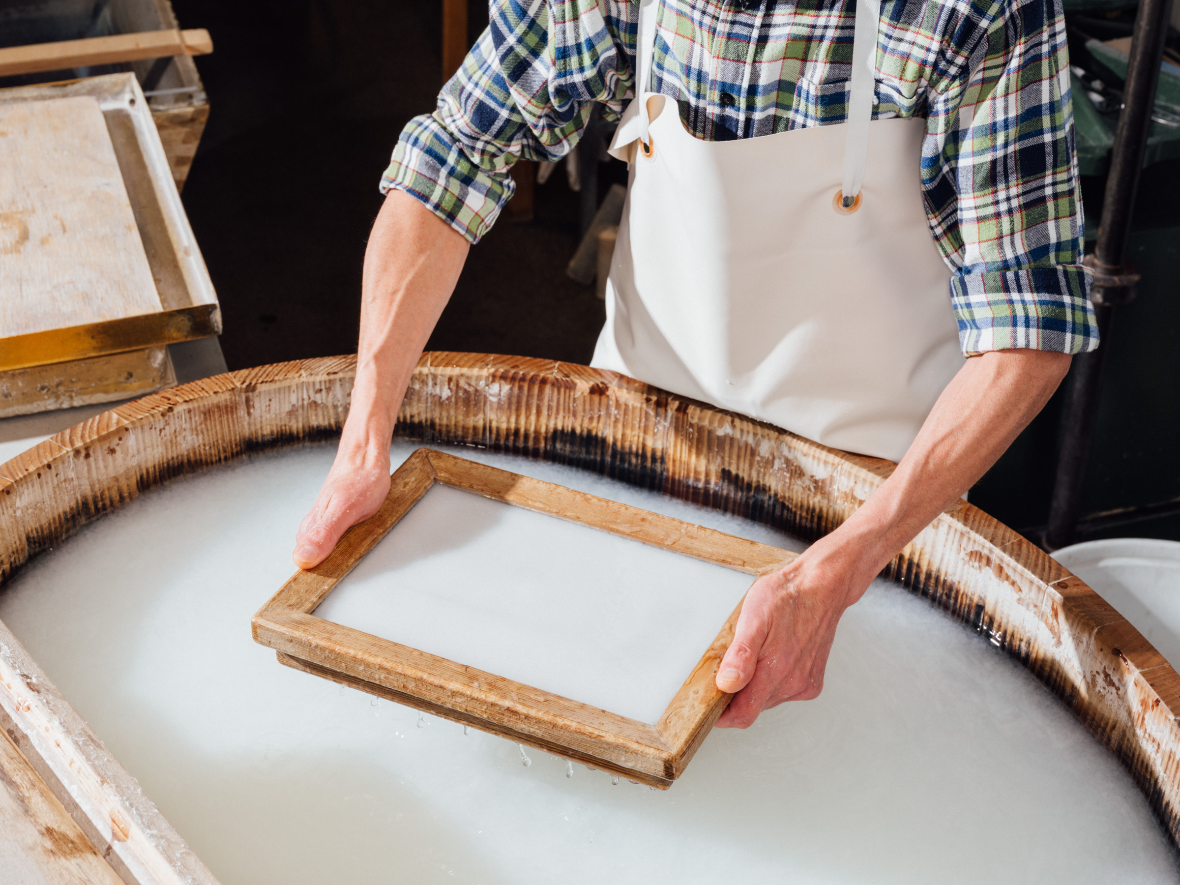 A man uses a sieve with a wooden rim to scoop paper from a vat of paper pulp.