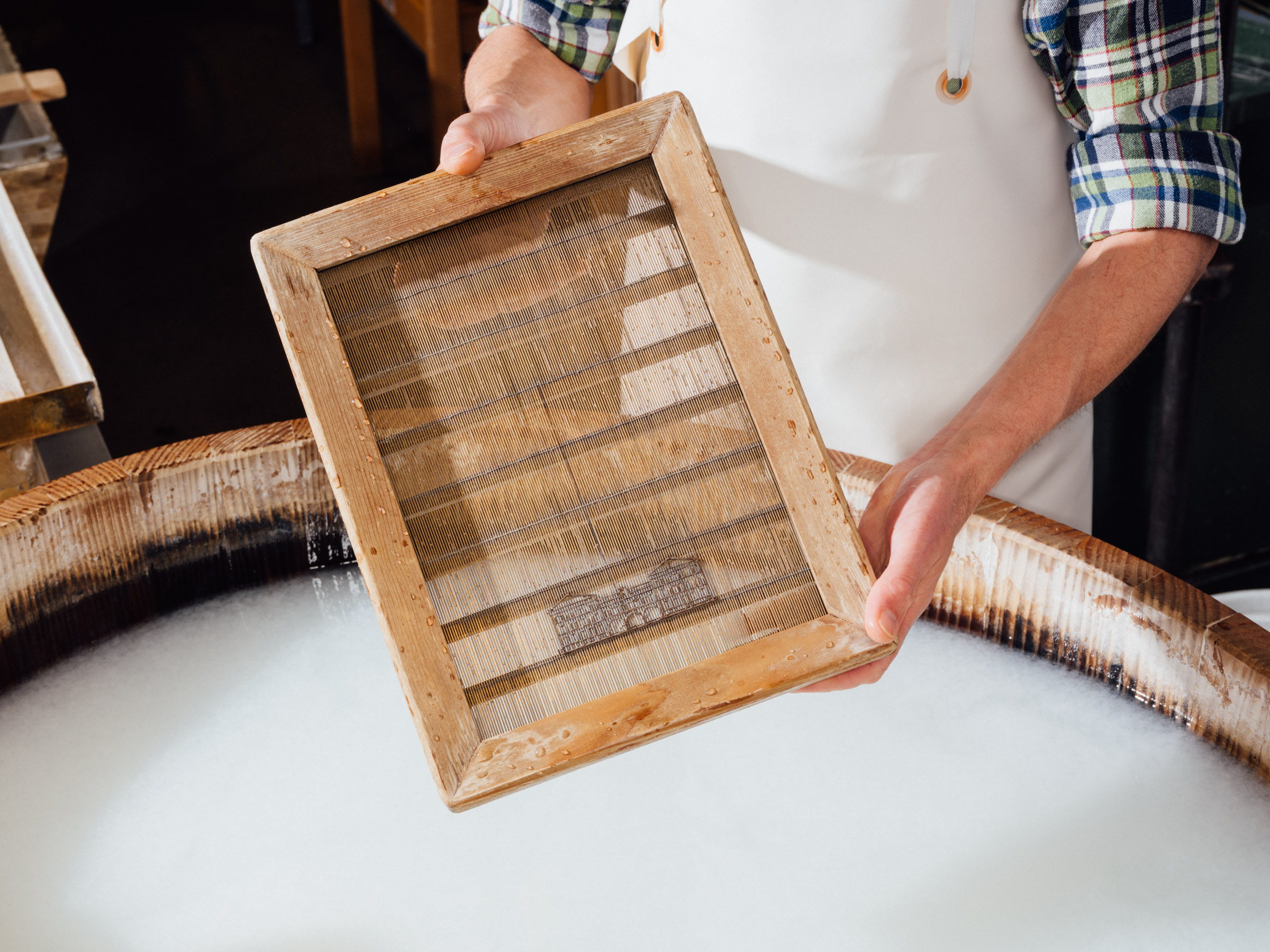 A man is holding a fine, square sieve with a wooden frame over a vat of milky-white liquid.