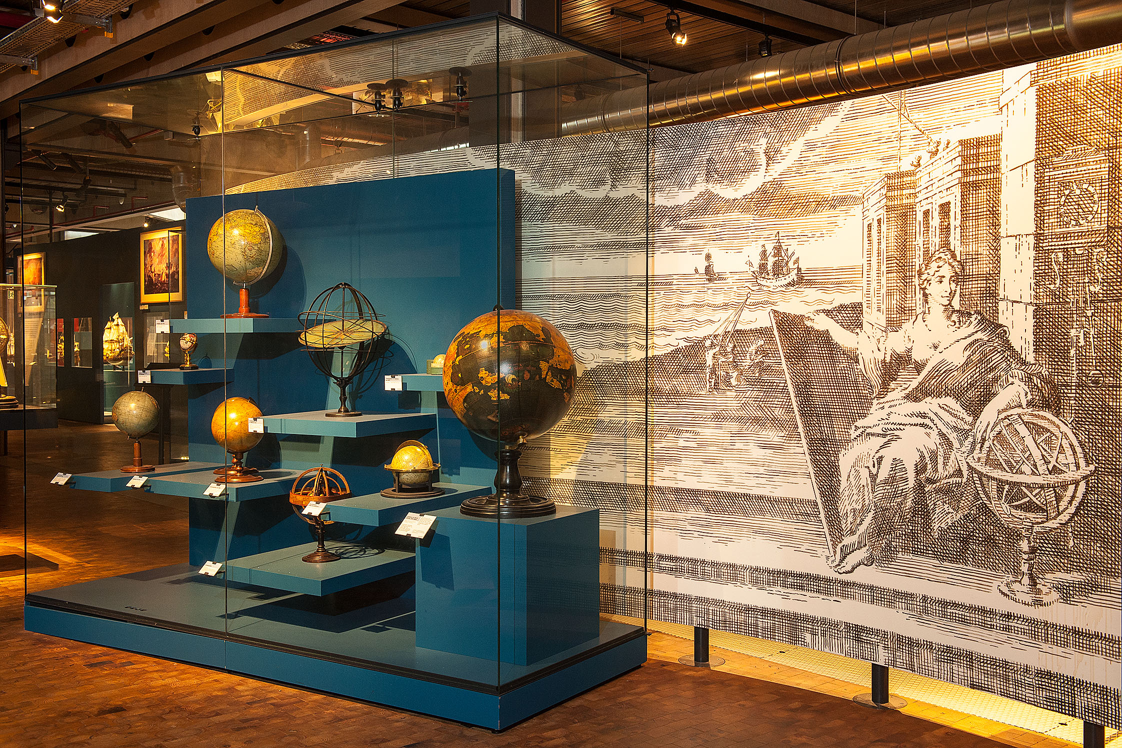  In front of a large black-and-white mural, various historical globes are displayed in a glass case with blue bases.