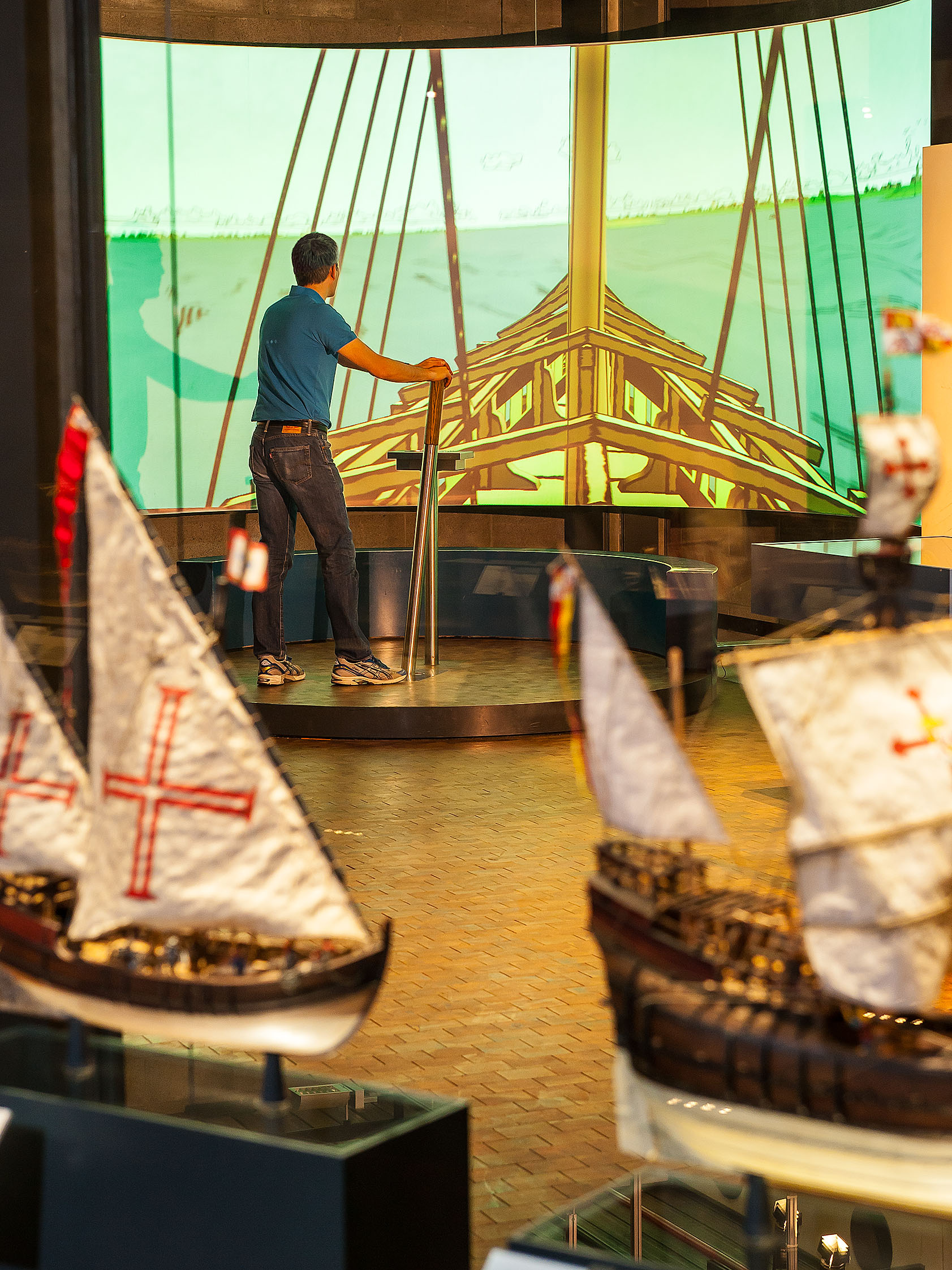 A man stands on a platform and steers a sailboat that is projected onto the wall in front of him.