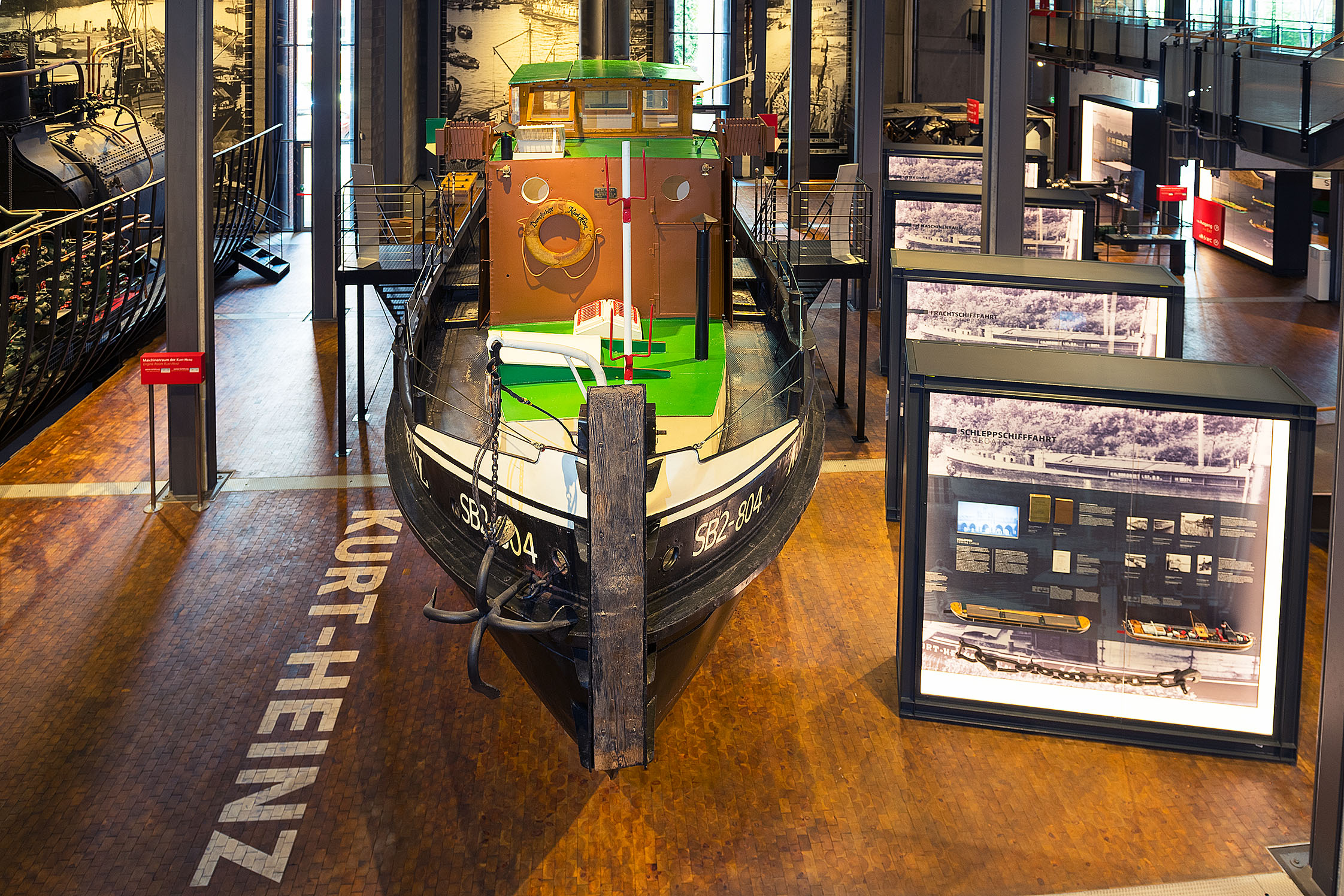 A small, historic steamboat is on display in the shipping exhibition. Next to it are large lighted display cases. On the side, the words “SB2-804” and “Kurt-Heinz” can be read. 