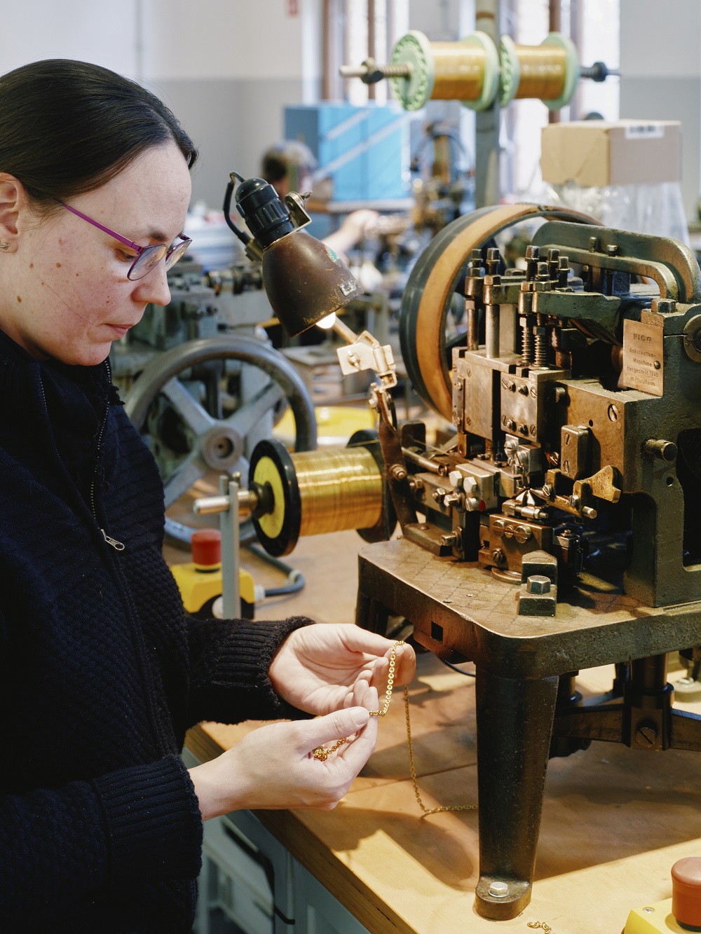 A woman is holding a gold necklace in her hand. In front of her is a machine used to produce these necklaces.