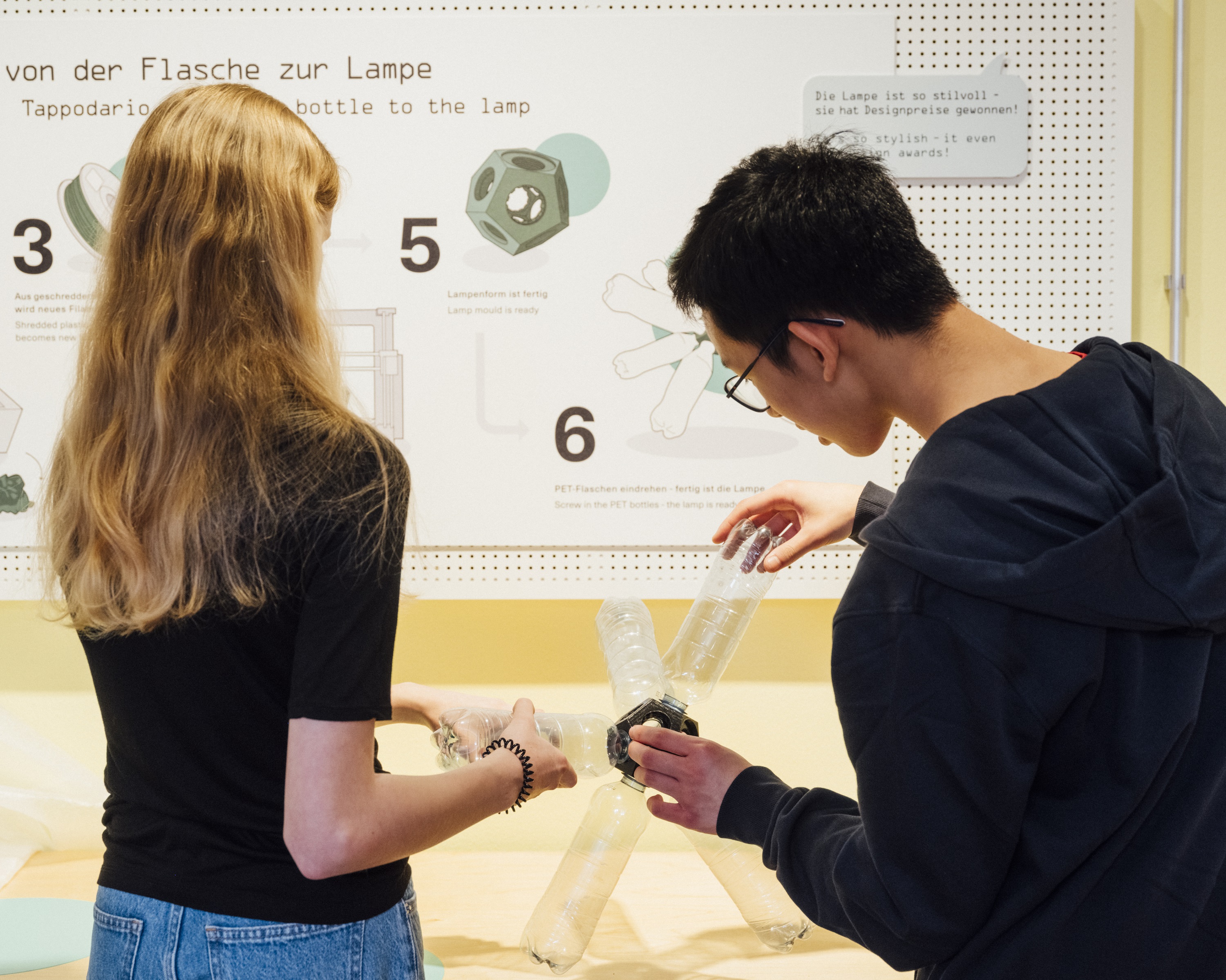 In front of a display panel with text and images bearing the caption “From Bottle to Lamp,” a girl and a boy are working together to screw empty plastic bottles into a plastic socket. The result is a lamp with a funny shape.