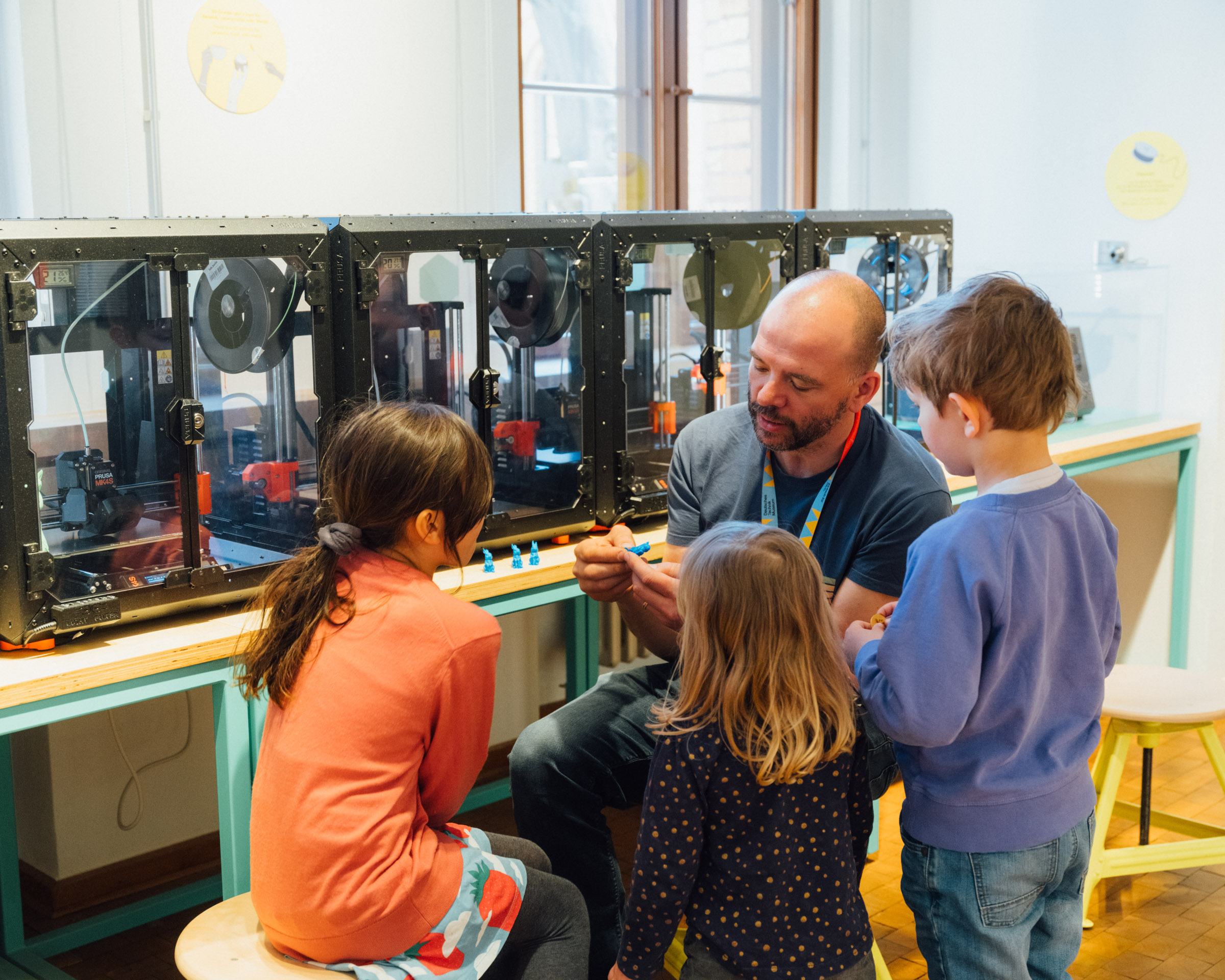 A man and three children are sitting in front of a row of 3D printers. The man is explaining something; the children are having fun and listening.