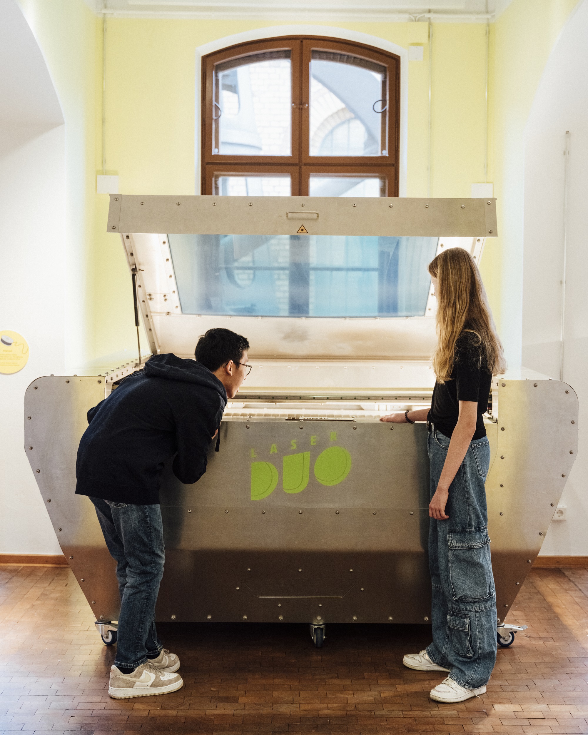 A girl and a boy are looking at a large, silver-colored piece of equipment. The word “LaserDuo” is written on the device. The boy is leaning over the opening of the device, and the girl is standing next to him, looking inside it.