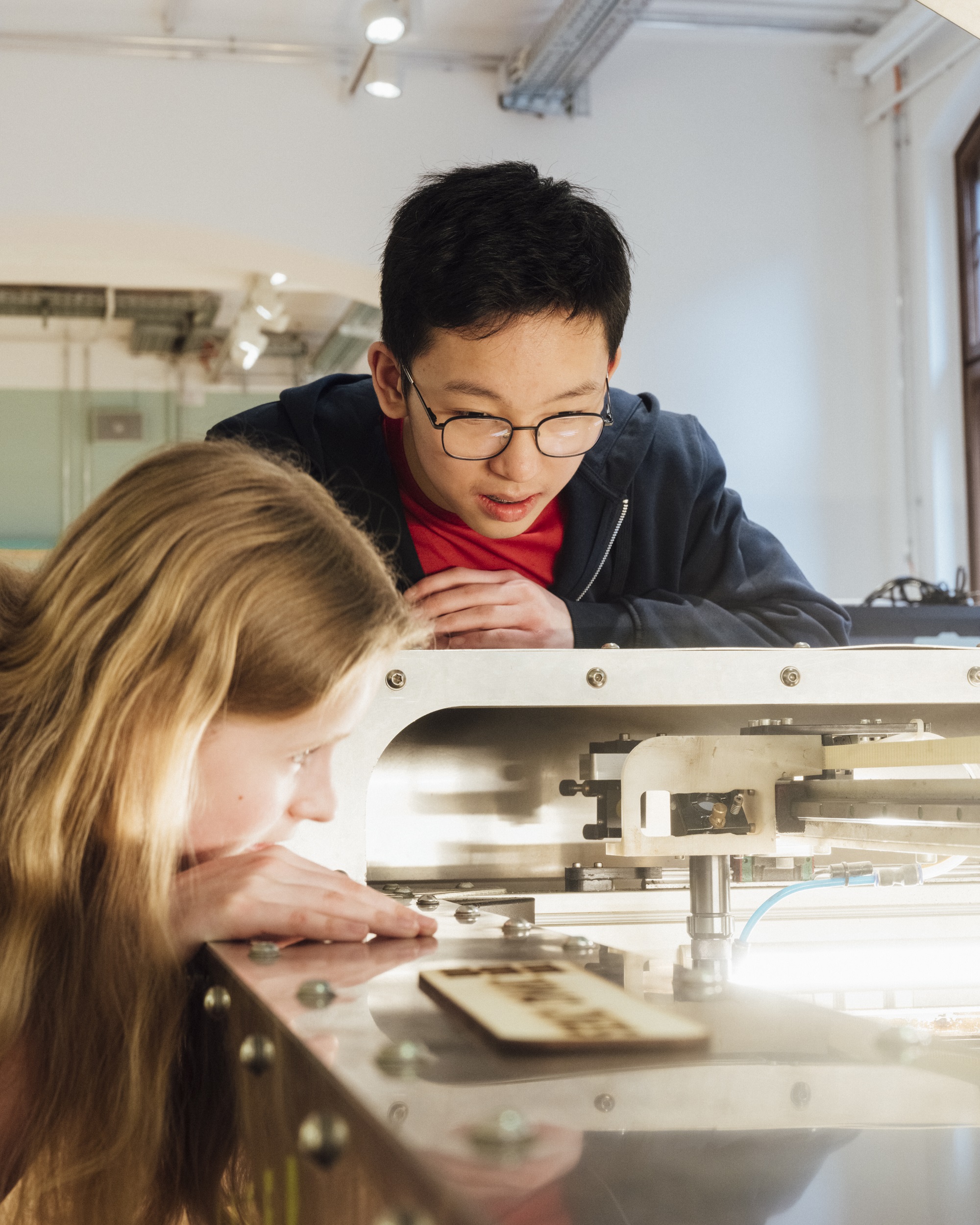 A girl and a boy are looking at a large piece of machinery. The girl is leaning over the opening to get a better look inside, while the boy is leaning against the edge of the machine.