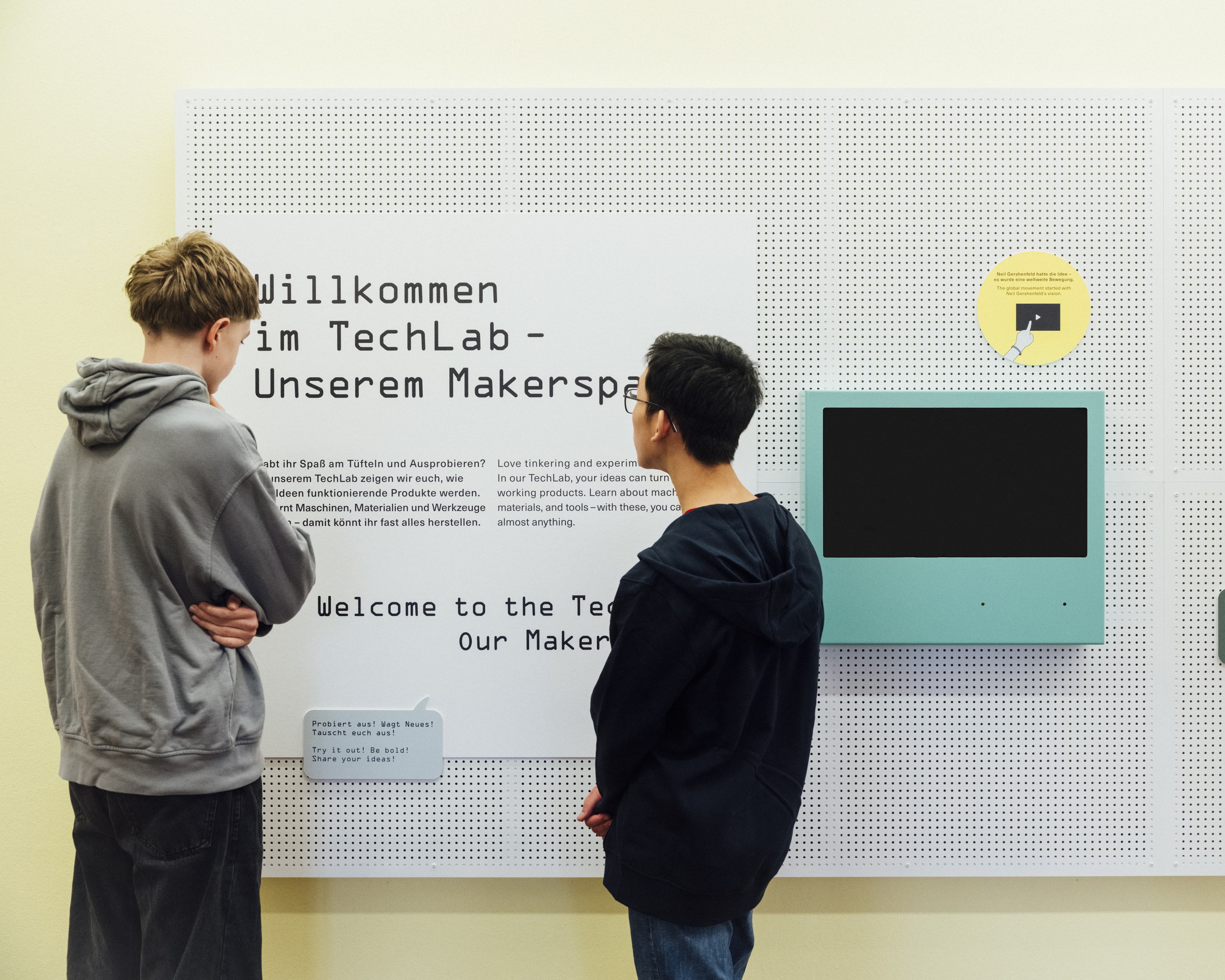 Two boys are standing in front of a sign that reads “Welcome to the TechLab—our makerspace” in German and English. They are reading the text. Next to it hangs a monitor with a blue border.