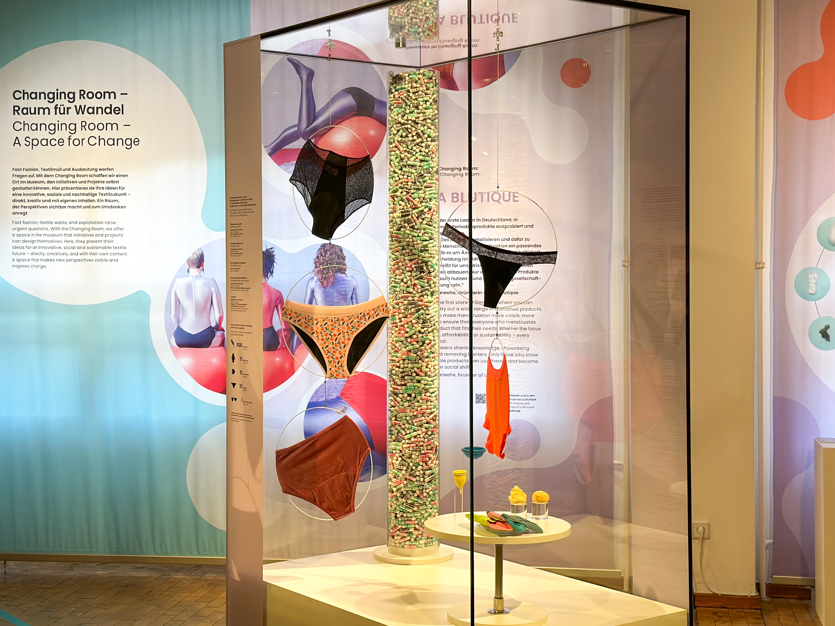 View of the display case in the "Changing Room" exhibition area: On display are various styles of period underwear, cloth pads, menstrual cups, and tampons. View of the display case in the "Changing Room" exhibition area: On display are various styles of period underwear, cloth pads, menstrual cups, and tampons. In the background, the words “Changing Room” can be seen.