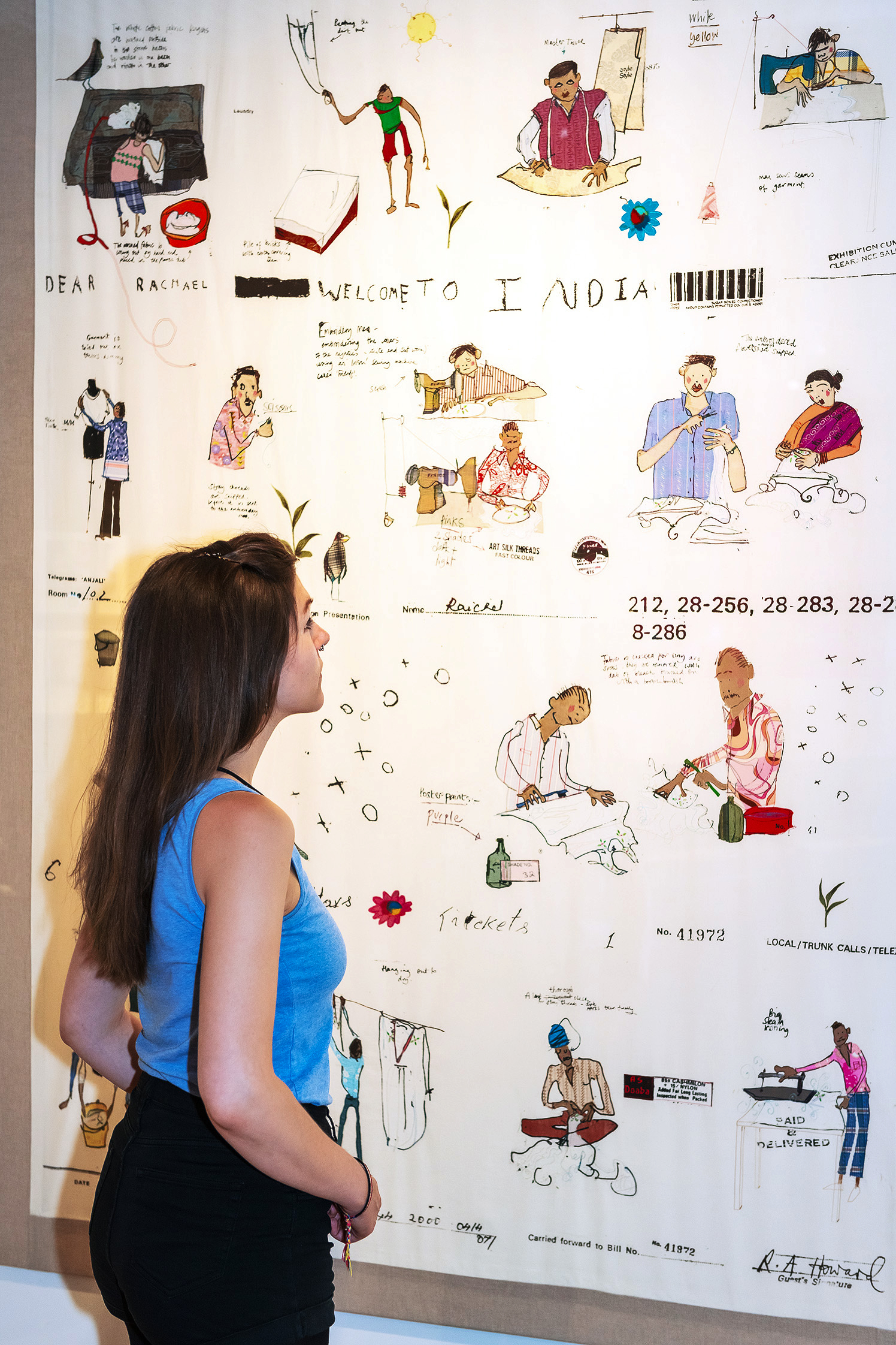 A young woman is looking at a white tapestry featuring various drawn figures engaged in different types of textile work. 