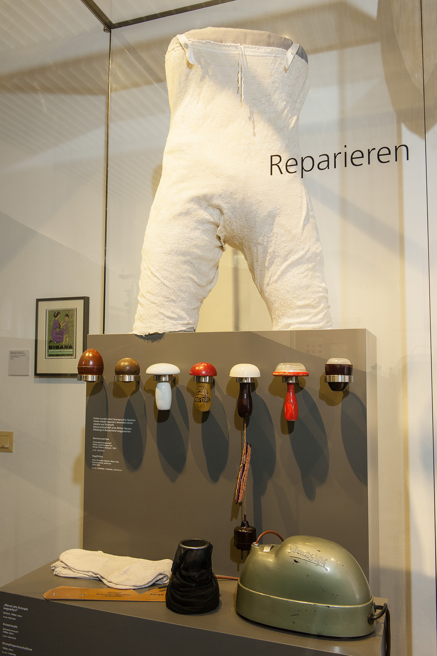 A pair of white underwear, heavily mended, sits on a pedestal inside a display case. Below the pedestal, various mending tools that resemble mushrooms are lined up side by side.