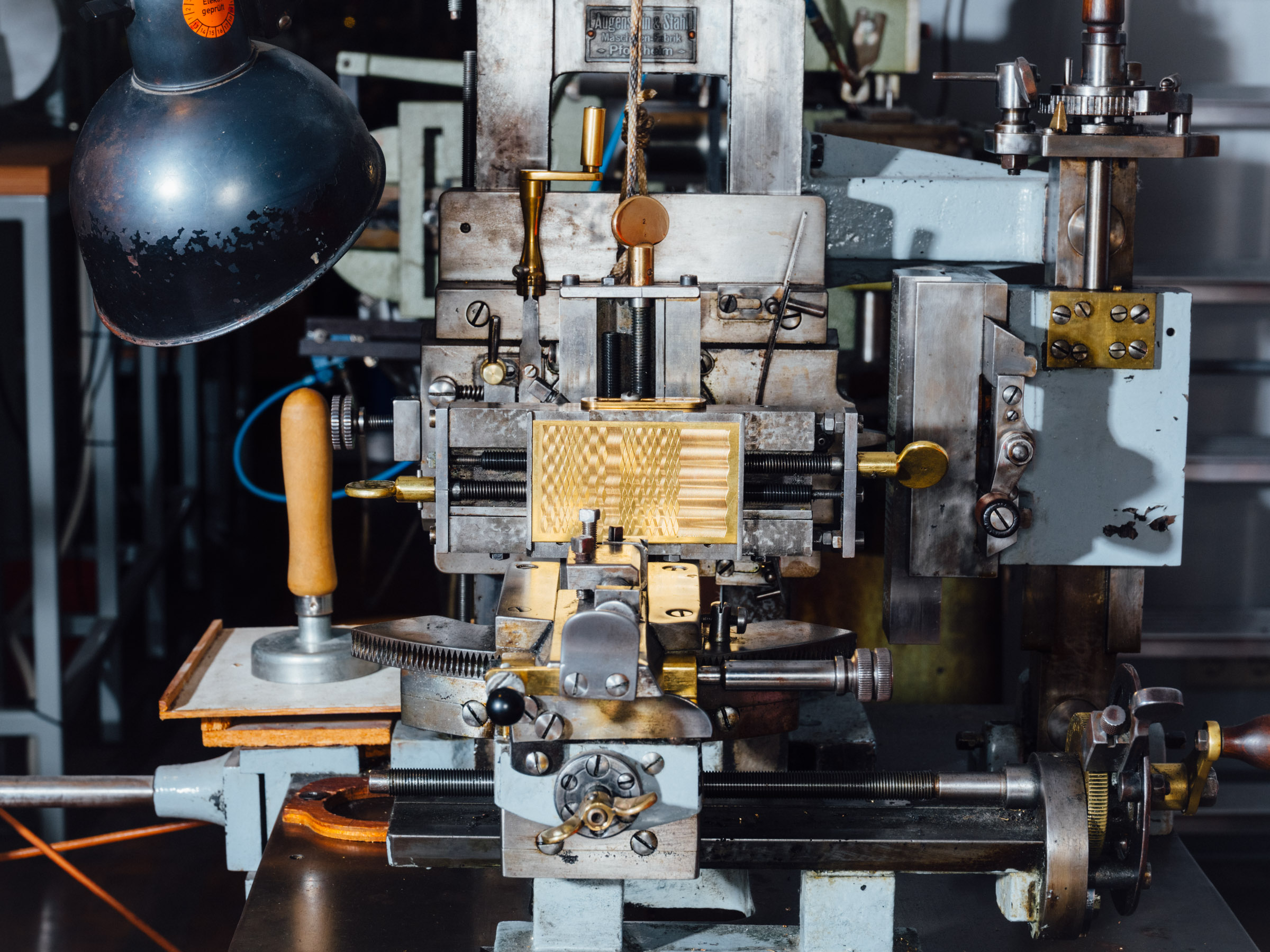 A metal machine with many levers; clamped in one place is a gold-colored piece of metal with geometric patterns engraved into it.