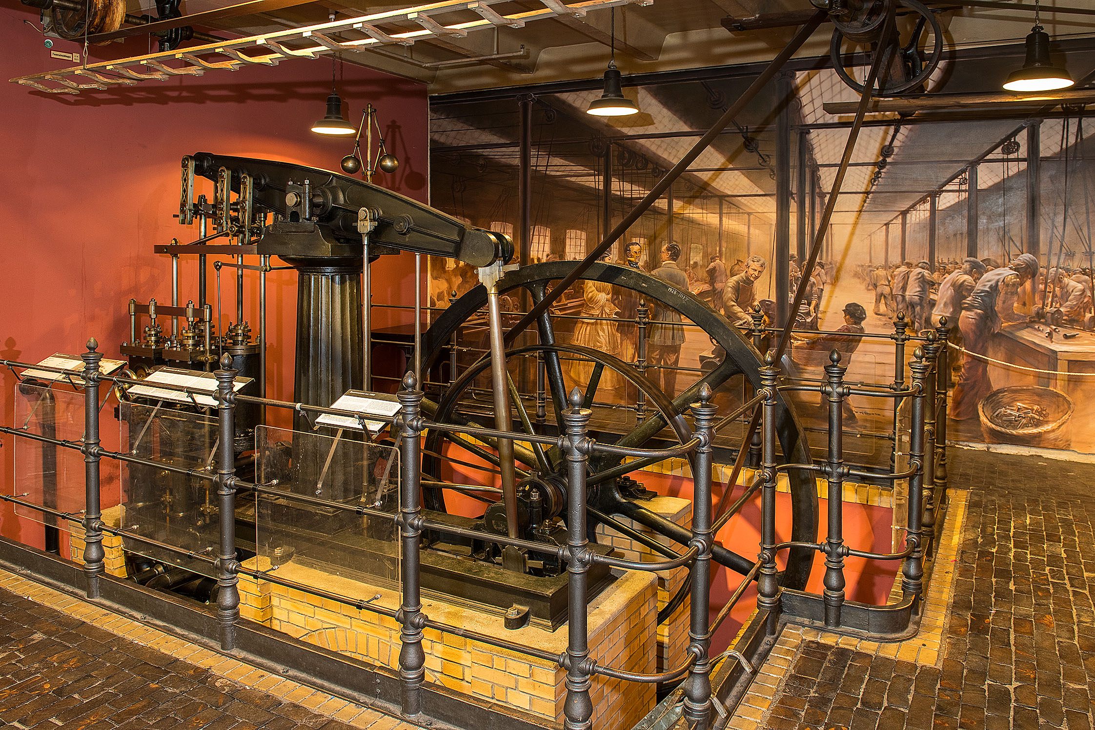 The central drive shaft is protected by a guardrail. You can see the smaller drive wheel with the drive belt and the larger flywheel behind it. In the foreground are the pistons of the steam engine; in the background is a mural depicting a historic workshop scene.
