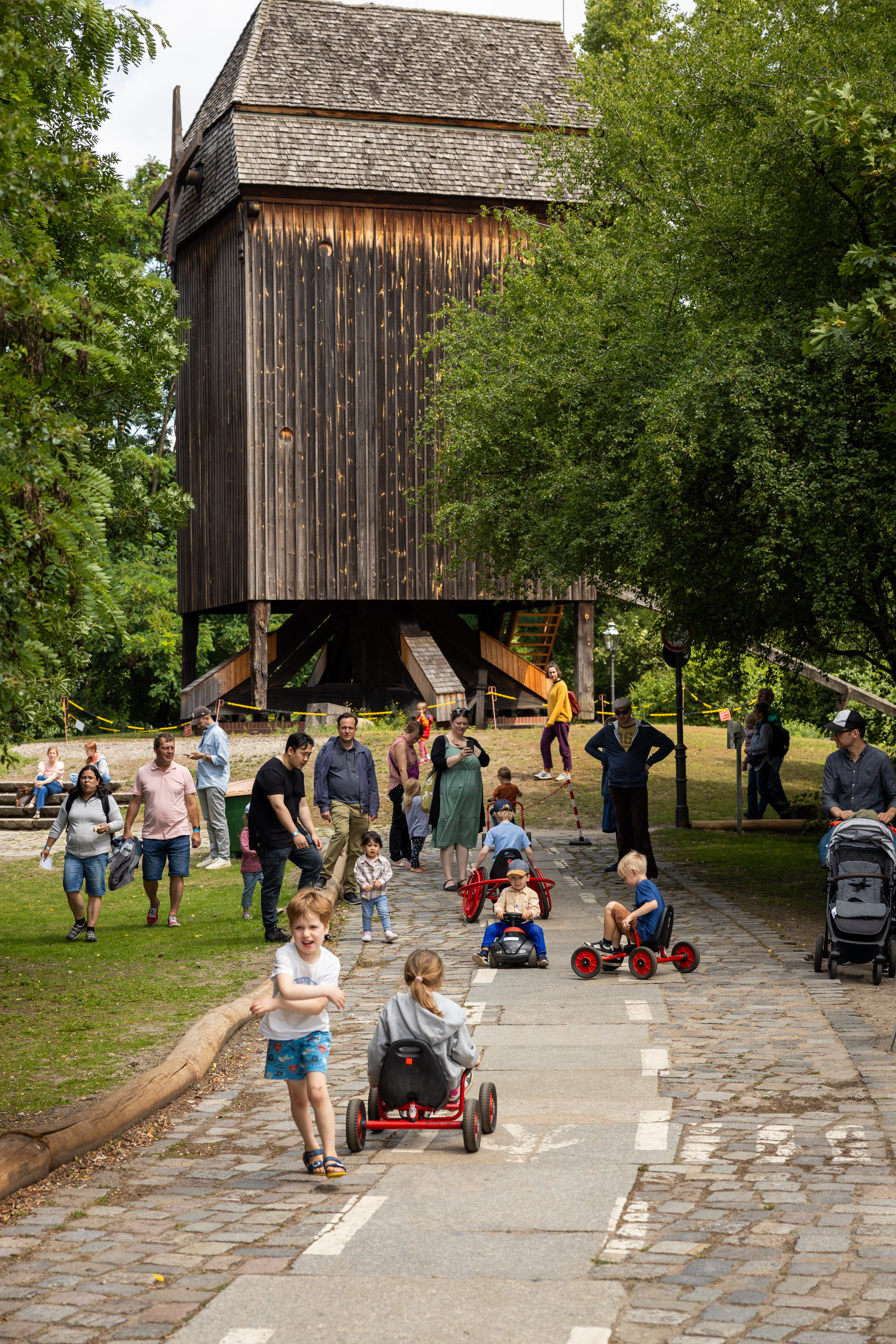 Kinder auf kleinen Fahruegen in einem Park, im Hintergrund eine Mühle