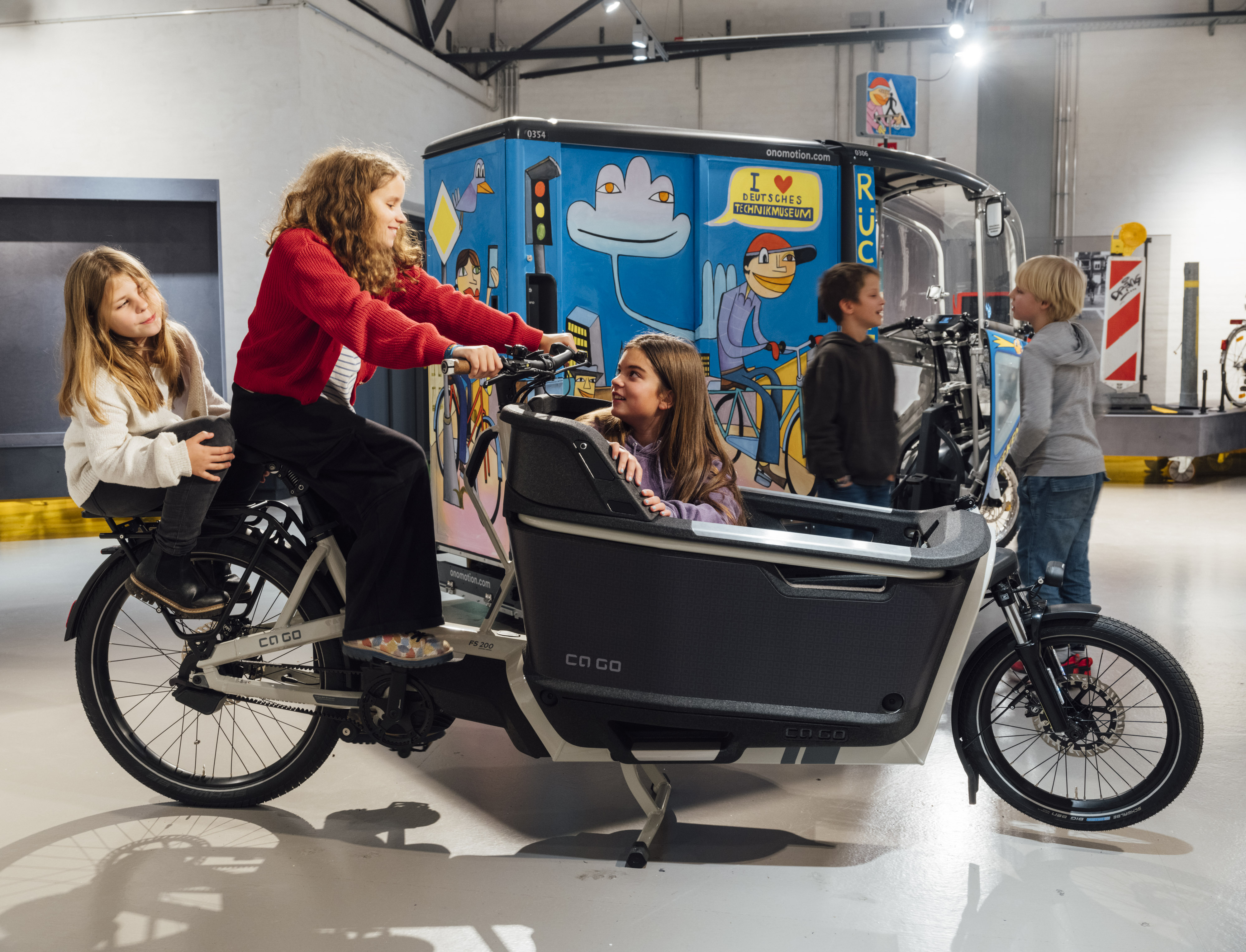 A view of an exhibition. Two girls are sitting on a modern cargo bike, and another girl is sitting in the cargo area. In the background, two boys are talking in front of a large, colorful electric cargo bike.