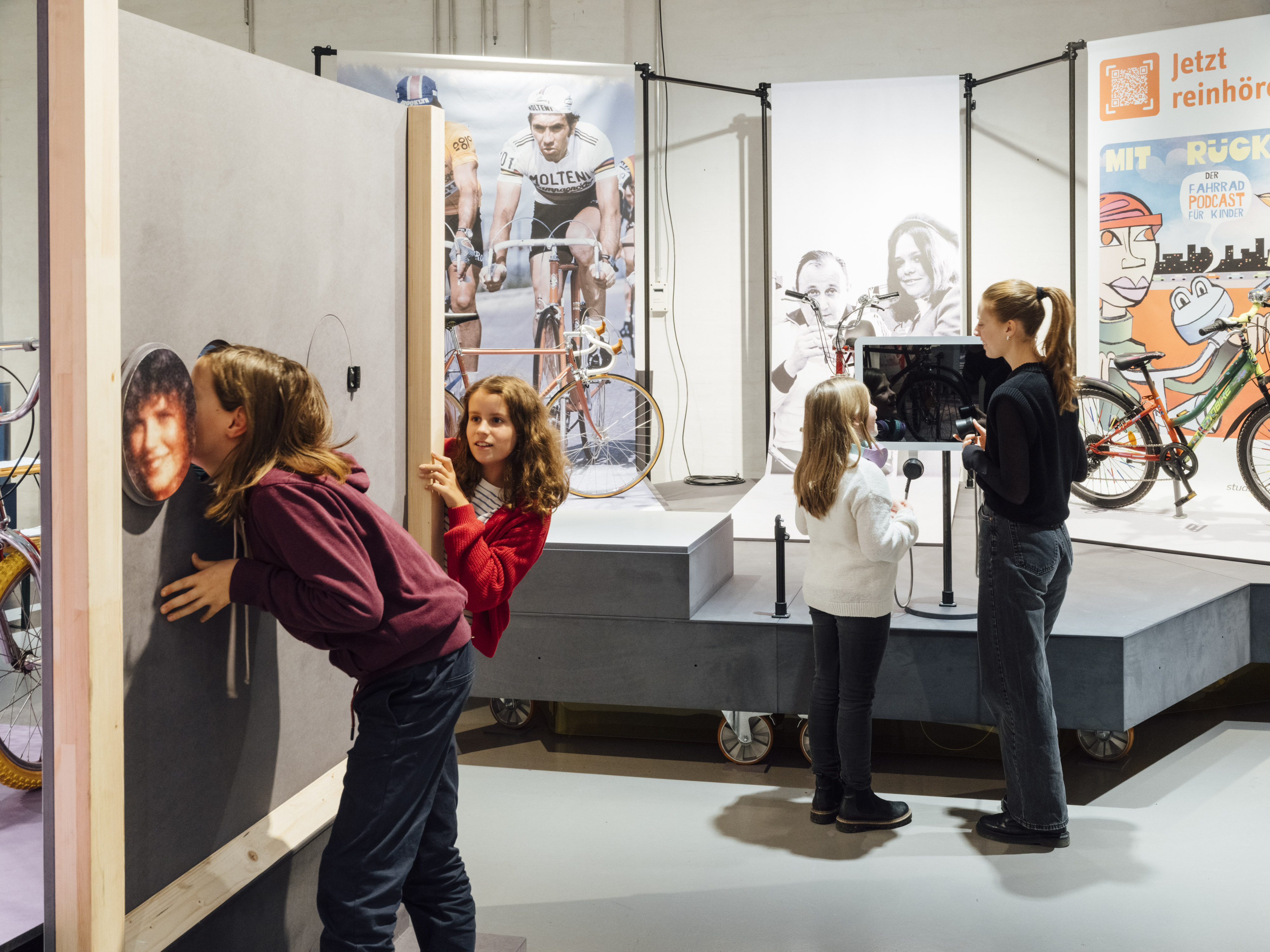 Several people in an exhibition hall. In the foreground, two young girls are examining a wooden wall with holes in it; in the background, a young woman and a little girl are talking in front of a display featuring bicycles and pictures of cyclists.