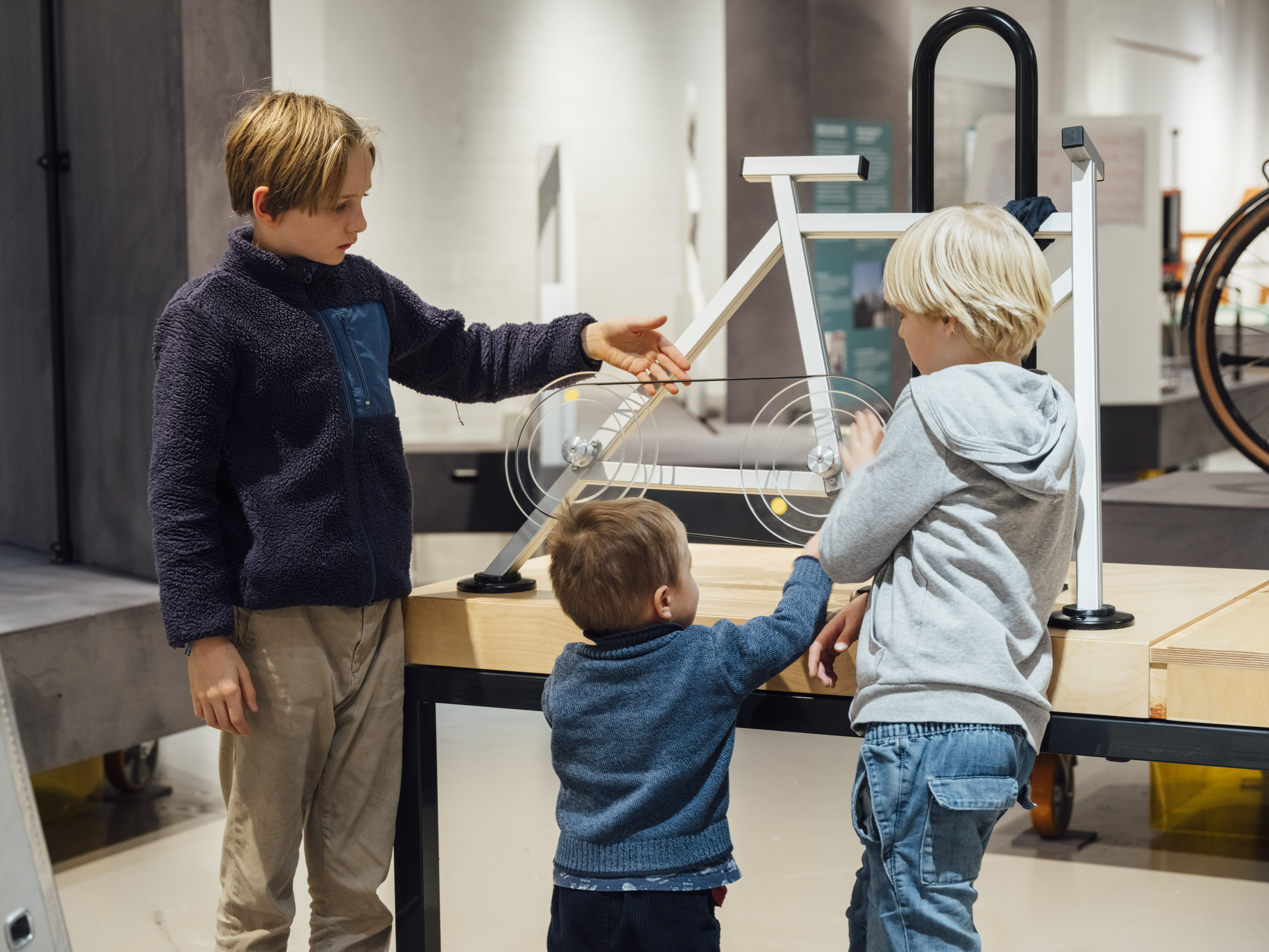 Three children are standing in an exhibition hall in front of a simplified, schematic replica of a bicycle. A boy is touching the bicycle chain depicted there.