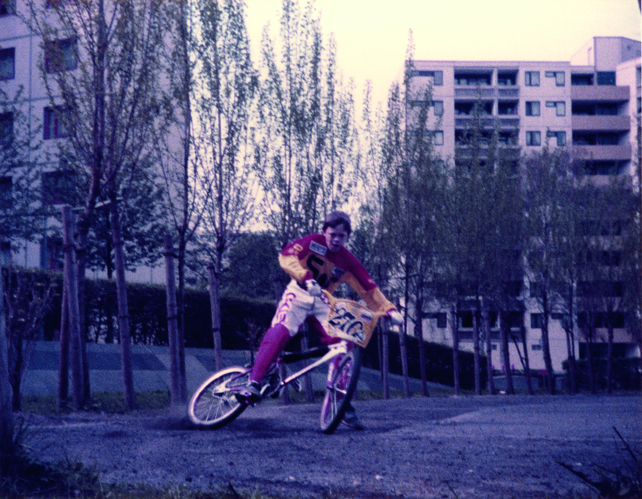 A child is riding a BMX bike against a backdrop of skyscrapers. He is braking and drifting to the side. The child is wearing BMX pants and a shirt, but no helmet; the BMX bike has a race number for sporting competitions attached to the handlebars.