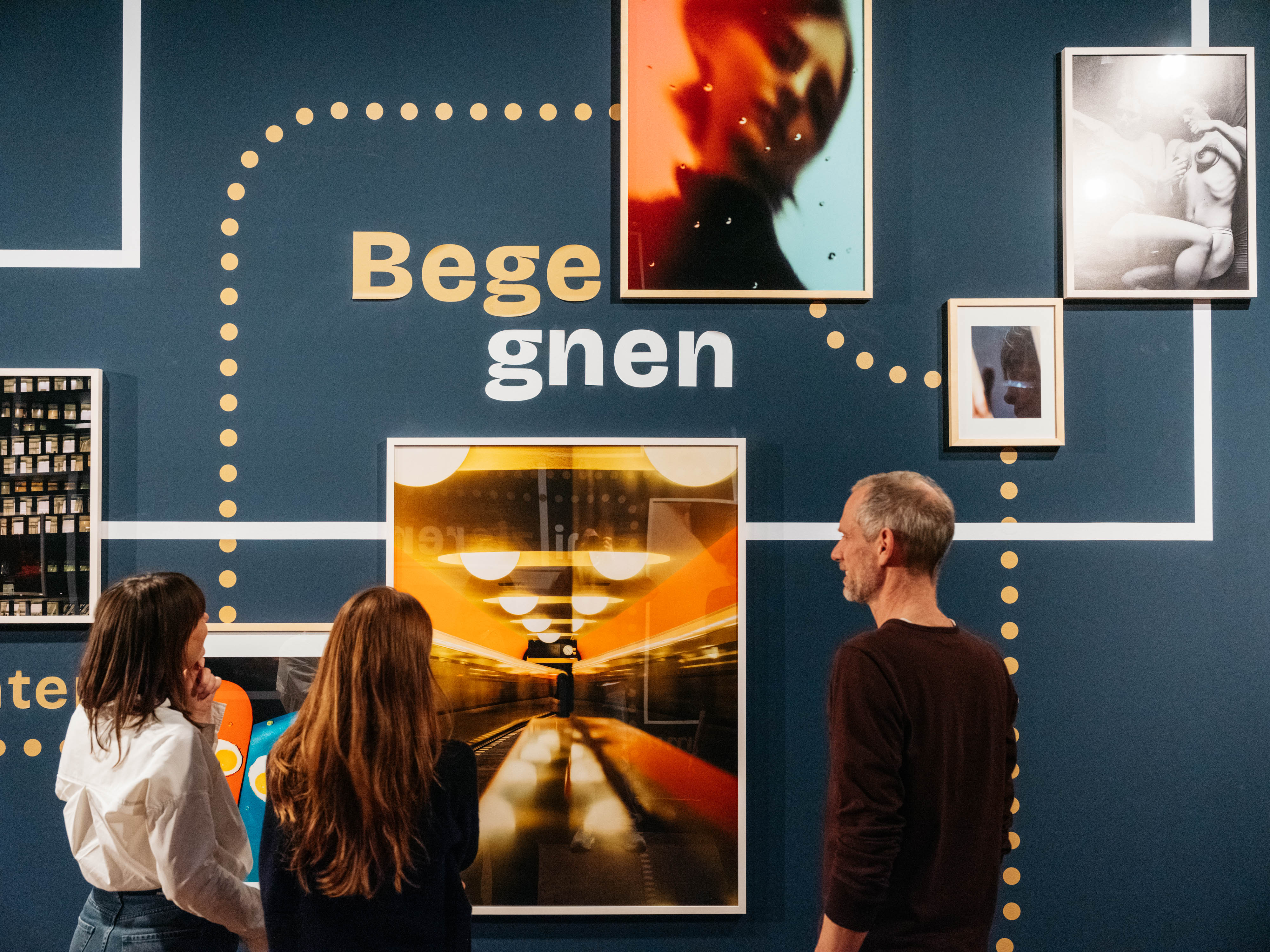 Several framed pictures hang on a dark blue wall. The word "Begegnen" is written in the center. Dotted lines and connecting lines can be seen between the individual photographs. Three people are standing in front of the pictures, looking at them.