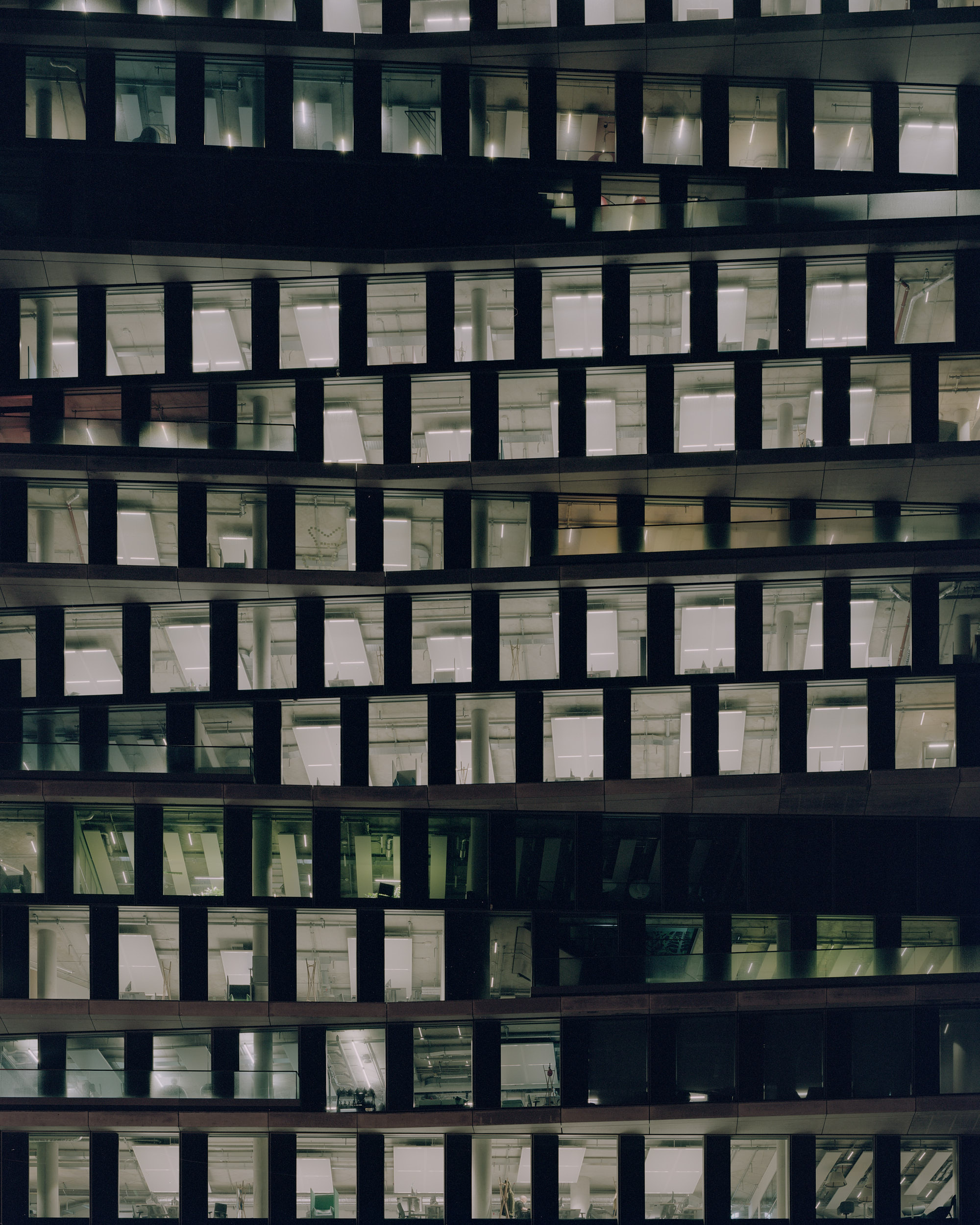 The color photograph shows a section of the facade of a modern office building at night. The windows are arranged irregularly, giving the structure a wavy appearance. Bright interior lighting illuminates the workspaces and technical installations. The dark exterior surfaces provide a stark contrast.