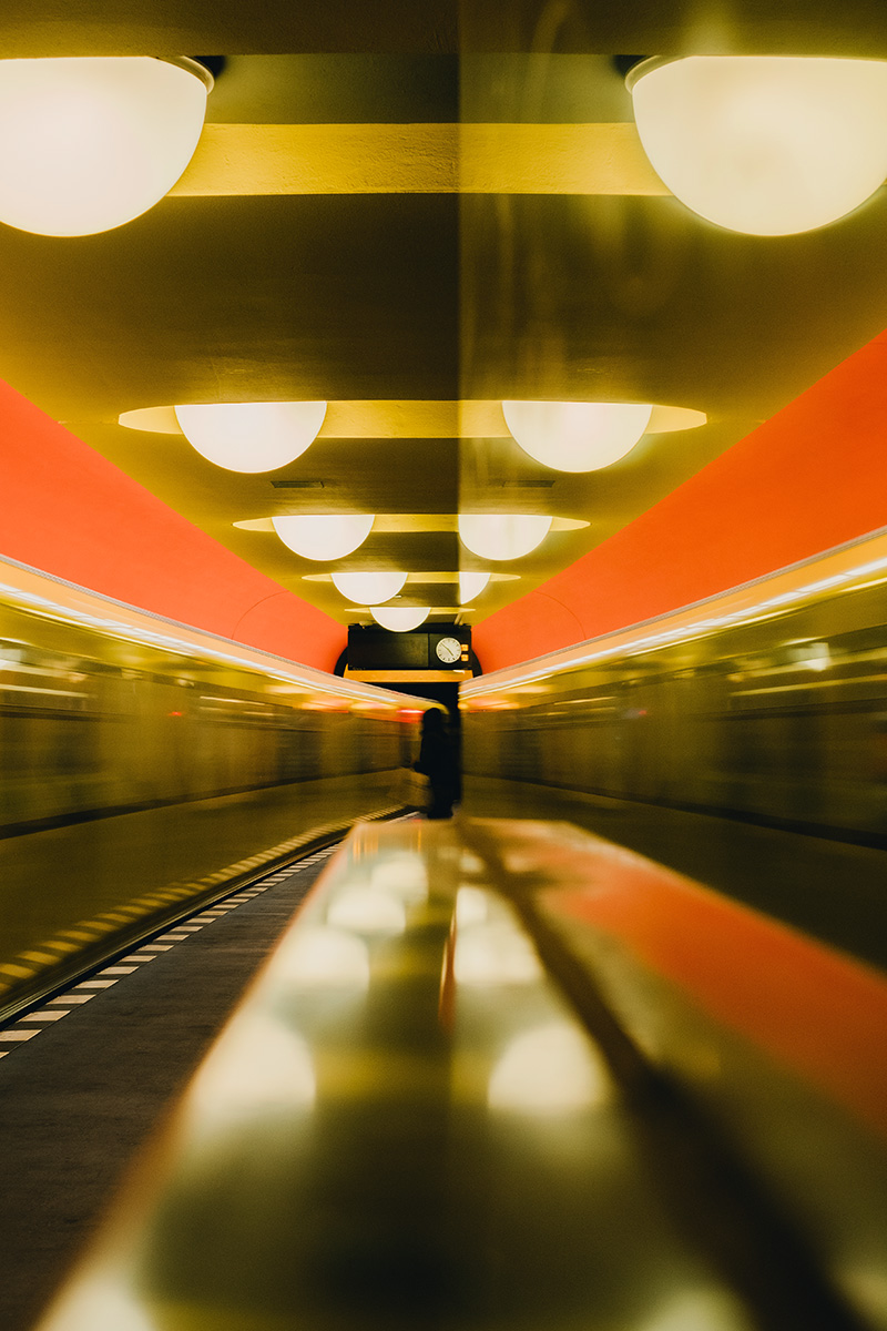 The color photograph shows a subway station with hemispherical ceiling lights. A train is just passing by on the left side. On the central platform, you can make out the silhouette of a person standing beneath the display board with a clock. The right side of the image is taken up by a surface on which the scene is reflected blurrily.
