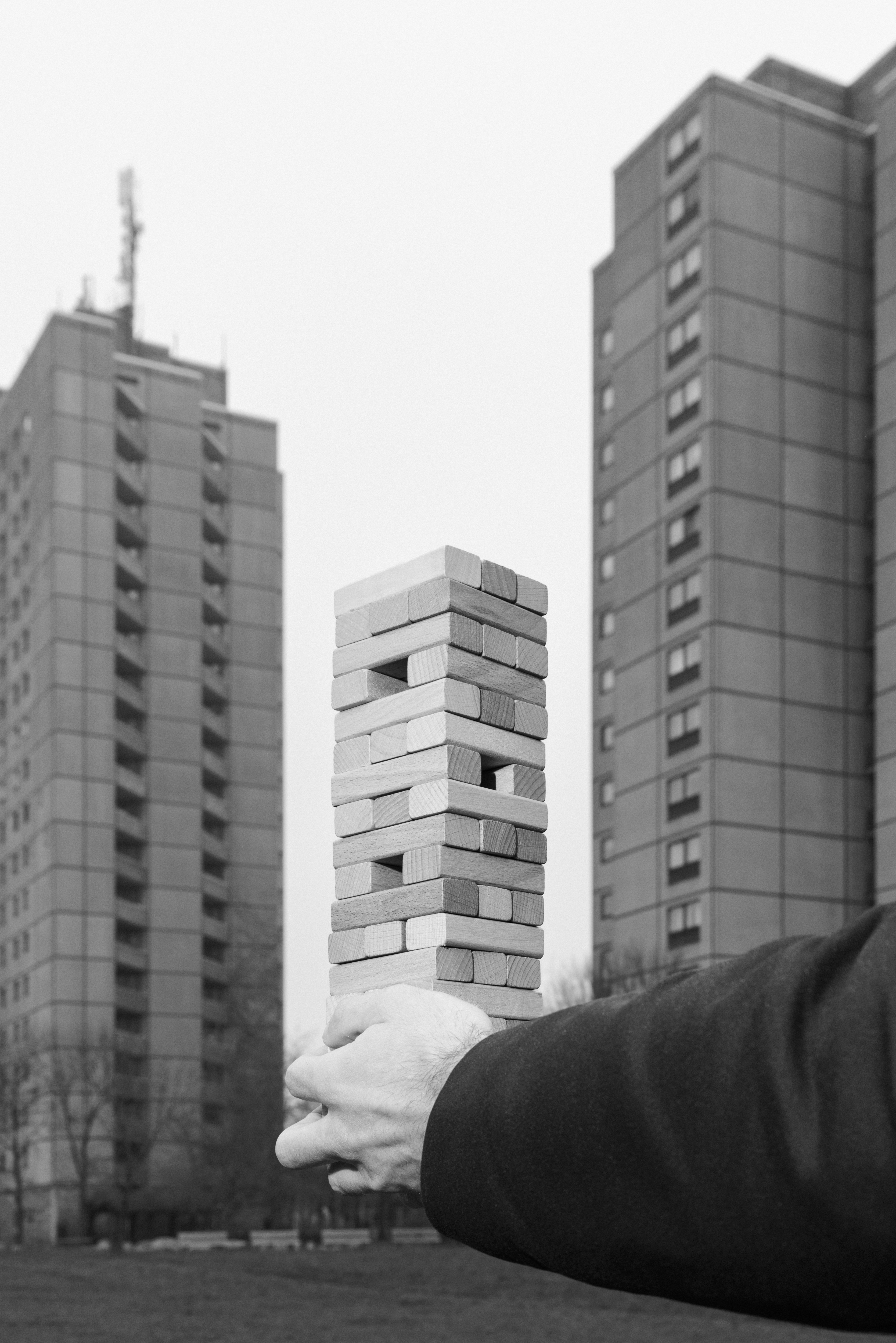 The black-and-white photo shows a hand holding a Jenga tower made of wooden blocks. In the background are two skyscrapers that visually echo the tower. Because of the perspective, it looks as if the Jenga tower were part of the architecture.