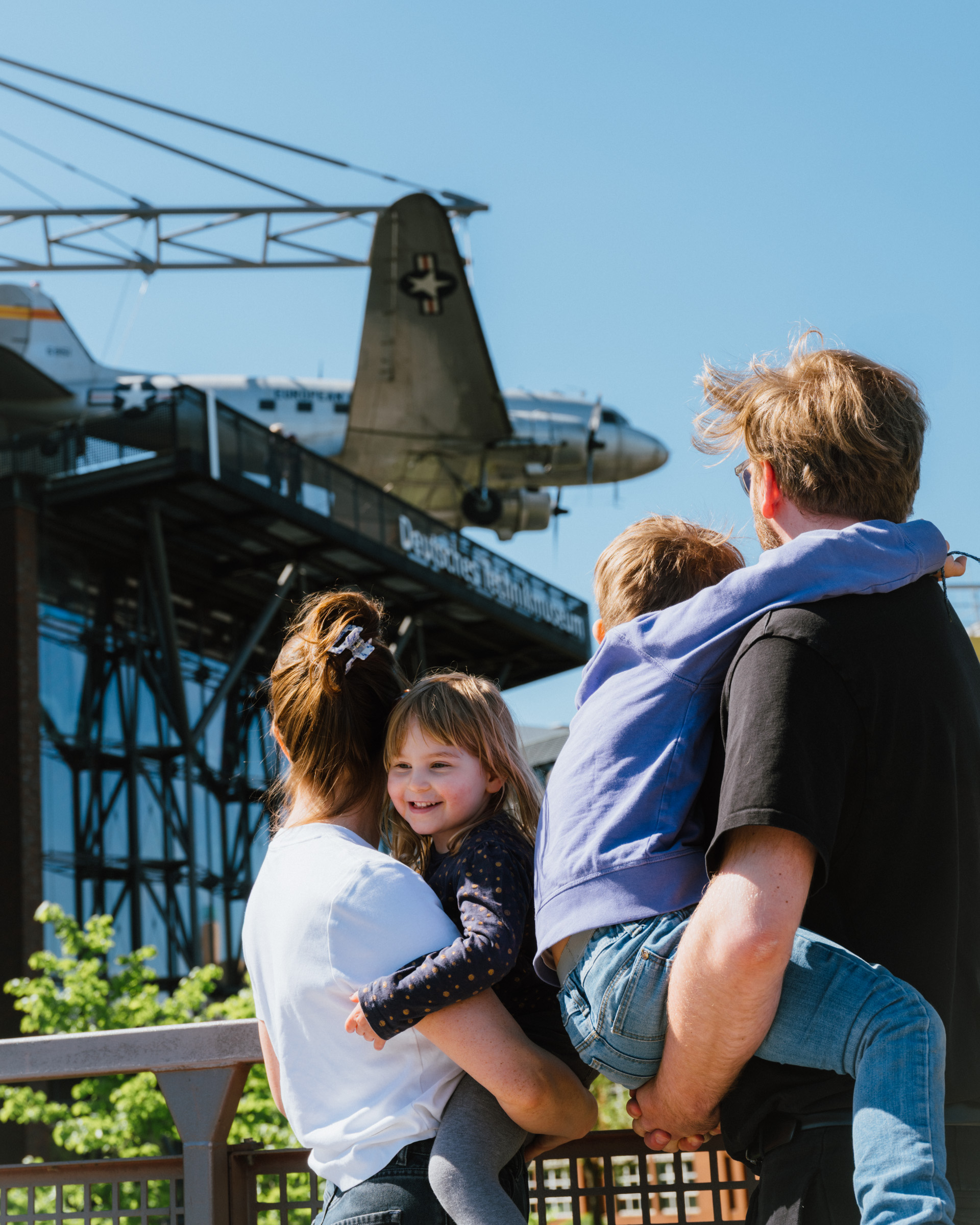 A couple with two children in their arms are standing on the walkway across from the museum, looking at the airplane hanging on the building's facade.